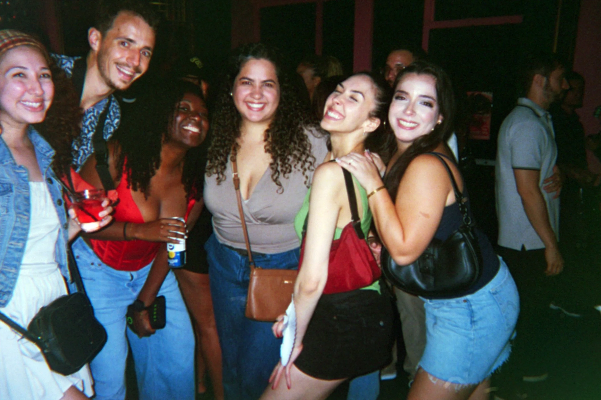 Group of six young women and one young man smiling and posing at a party, with some holding drinks, in an indoor setting with dark background.