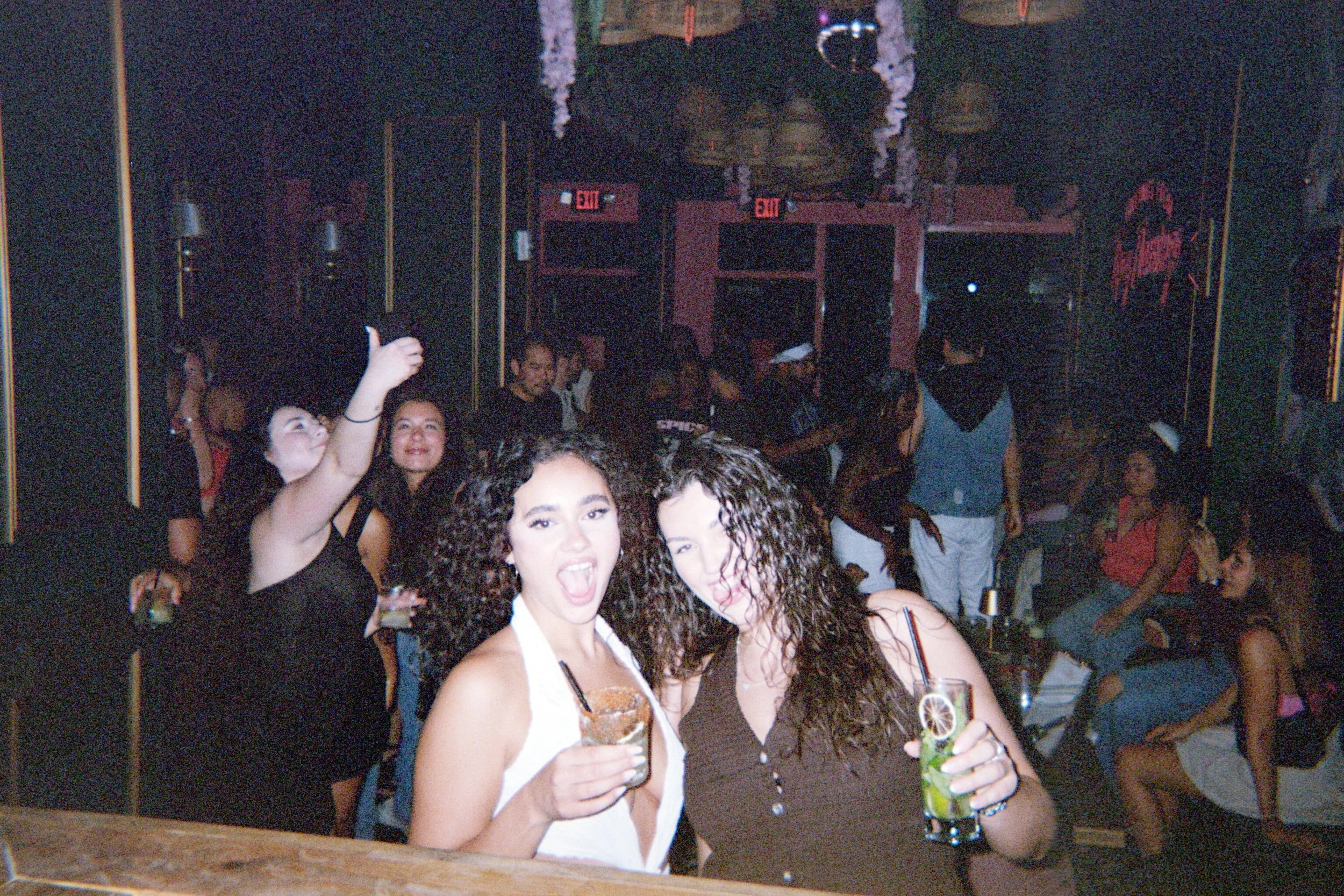 Two women with curly hair enjoying drinks at a crowded nightclub or bar, with other people dancing and socializing in the background.