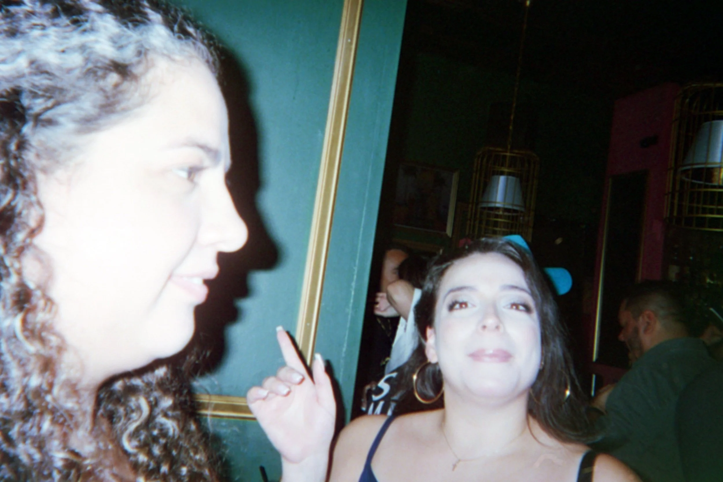 Two women, one with curly hair and the other with dark straight hair, smiling at each other in a dimly lit indoor setting.