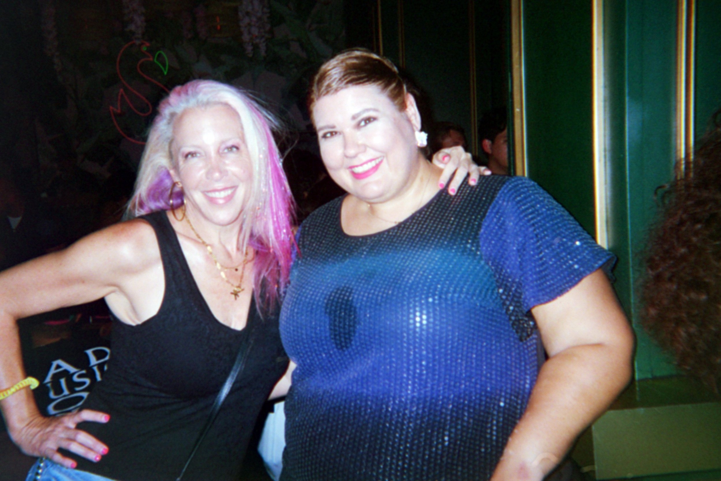 Two women posing together at a social event, smiling at the camera. One has long blonde hair with pink highlights, wearing a black sleeveless top with layered necklaces. The other has short brown hair, wearing a sparkling blue top, with her arm aroun