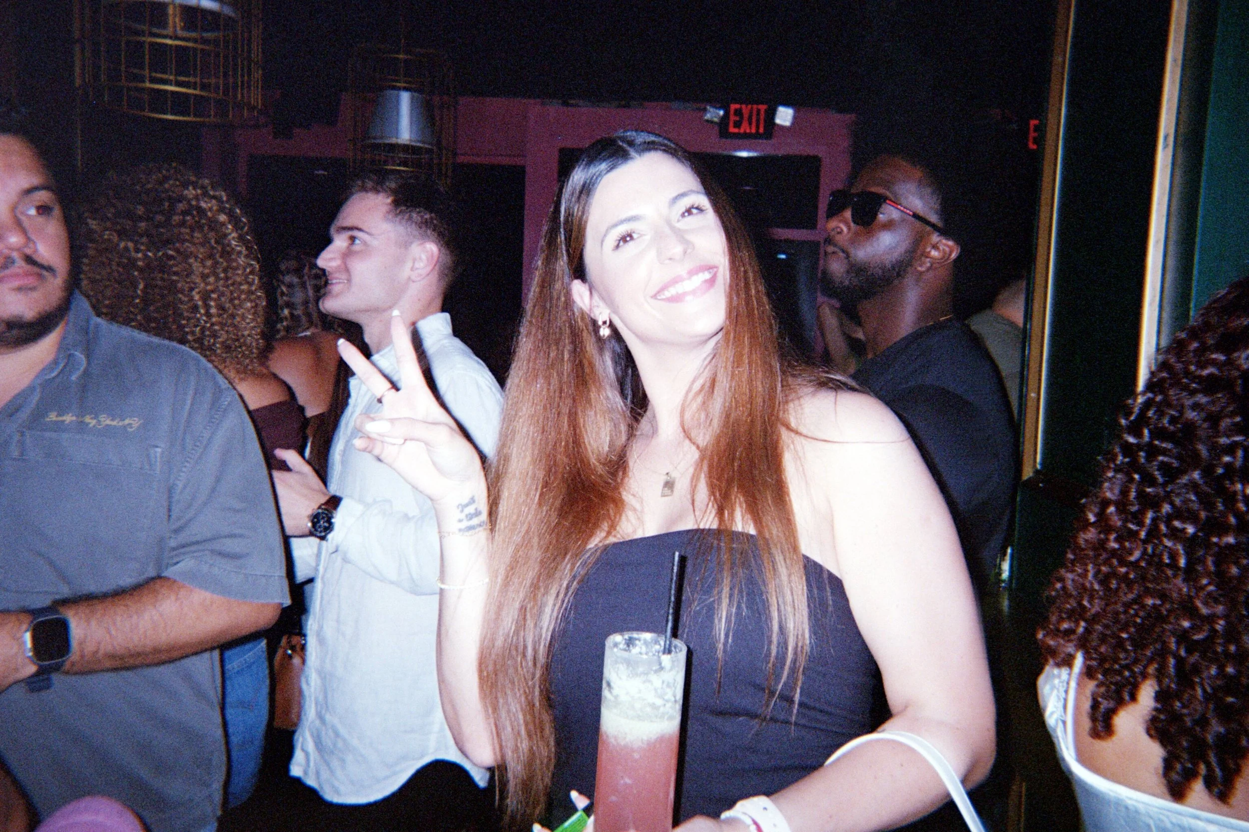 A young woman with long red hair smiling and making a peace sign, holding a colorful cocktail in a nightclub or bar setting with other people around her.