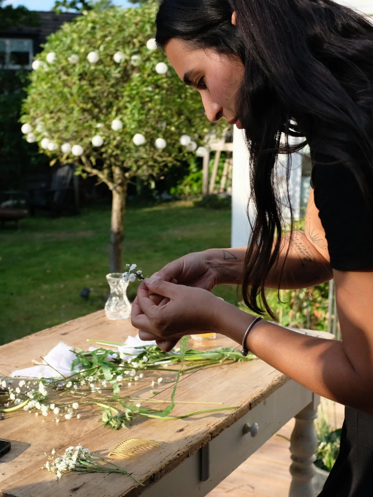 Woman arranging small white flowers on a wooden table outdoors, with a tree and lawn in the background.