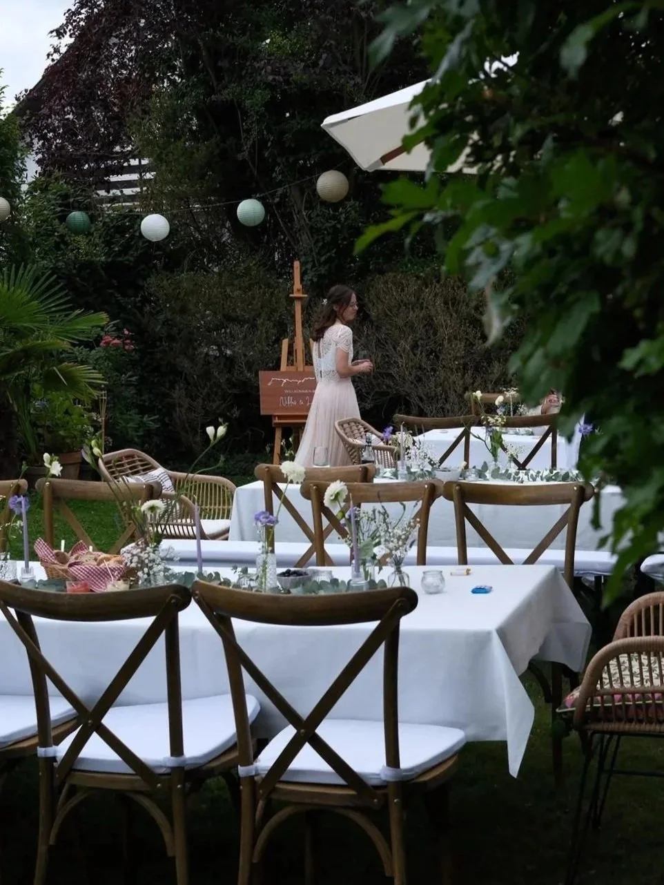 Outdoor evening event with white-clothed tables decorated with small vases of flowers, surrounded by wooden chairs, set in a garden with string lights and greenery. A woman in a white dress stands near a chalkboard artist's easel.
