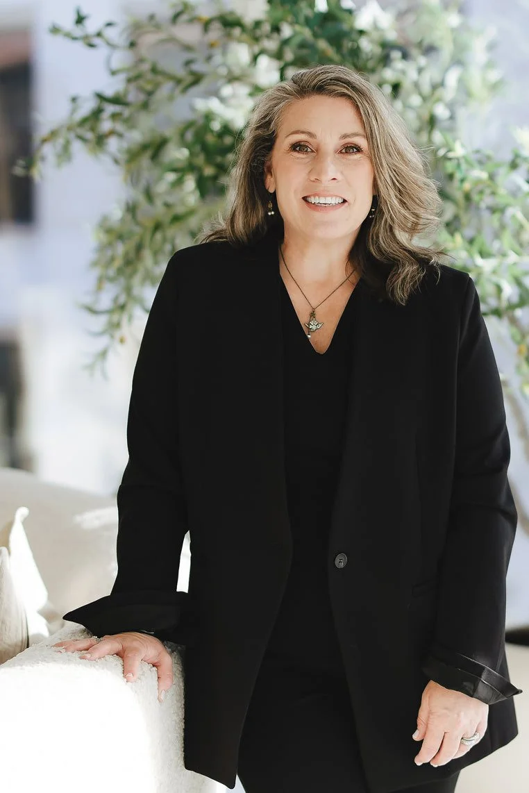 A woman with shoulder-length red hair, wearing hoop earrings, a black sleeveless turtleneck top, a black leather skirt with a side slit, a watch, and a ring, stands with her hand on her hip in a bright, spacious room with large windows and blurred background furniture and decor.