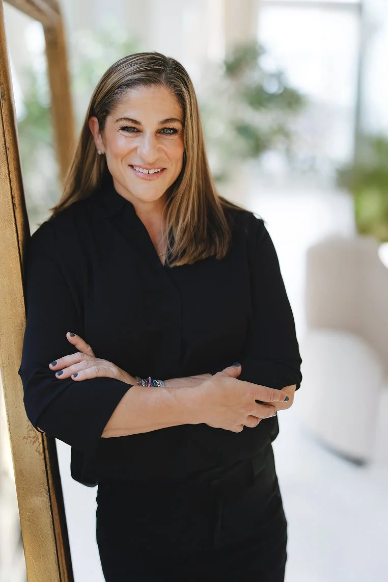 A woman with shoulder-length brown hair wearing a sleeveless black top and a black leather skirt standing indoors in front of large windows, with her left hand on her hip.