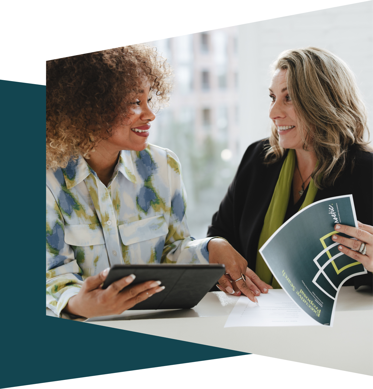 Two women smiling and discussing documents at a table inside an office, with one holding a tablet and the other holding a brochure.