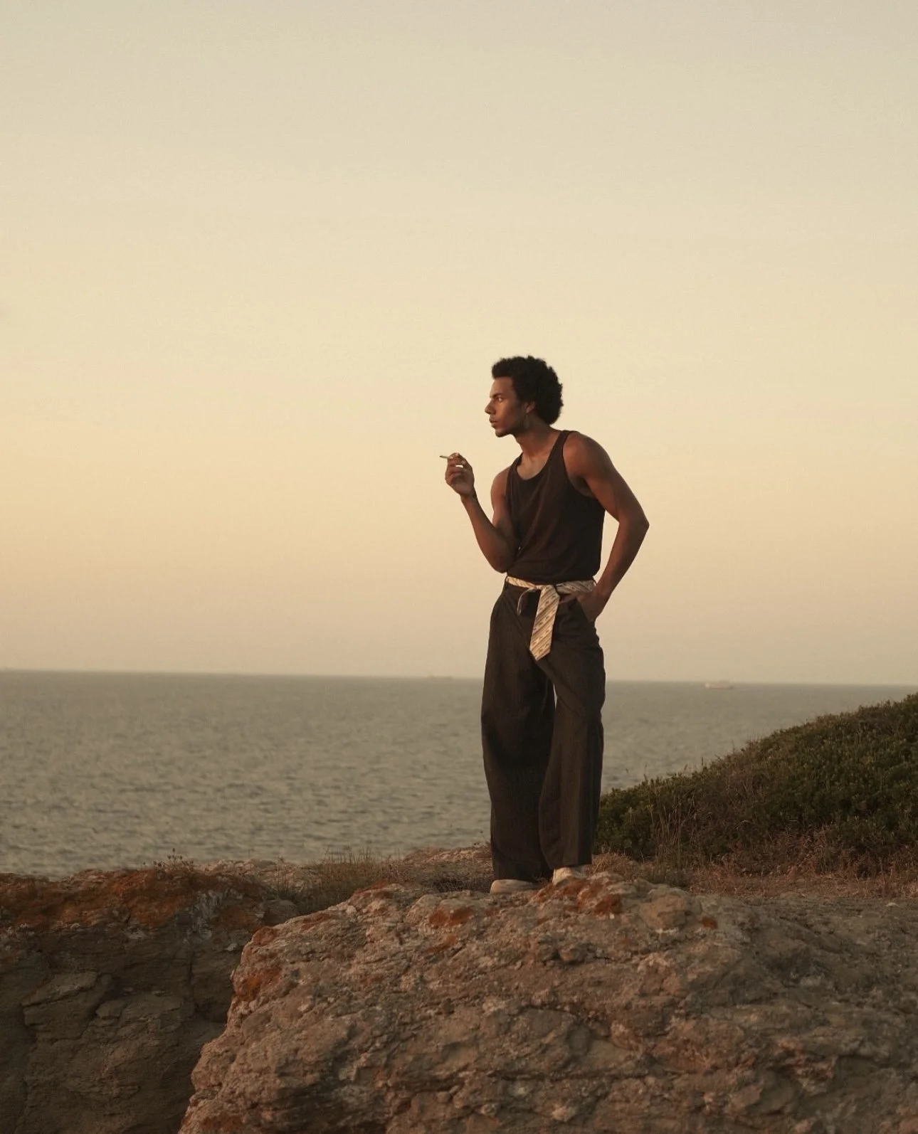 A person with curly hair standing on a rocky shoreline, holding a cigarette, and looking sideways towards the ocean during sunset.