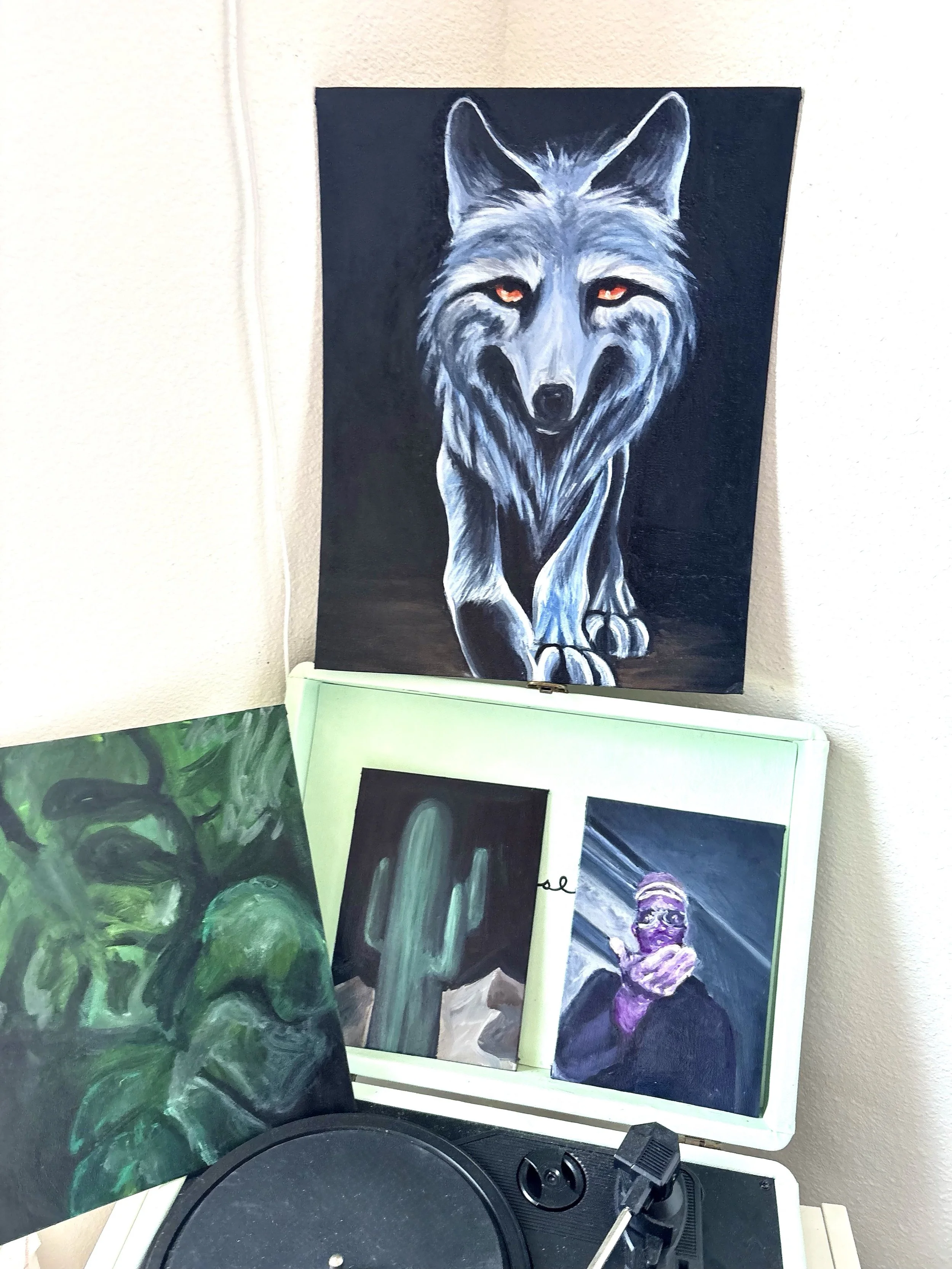 Collection of four paintings, including a large portrait of a wolf with red eyes on the wall, and three smaller paintings on a table featuring a green face, a cactus, and a person with purple face paint and a face covering.
