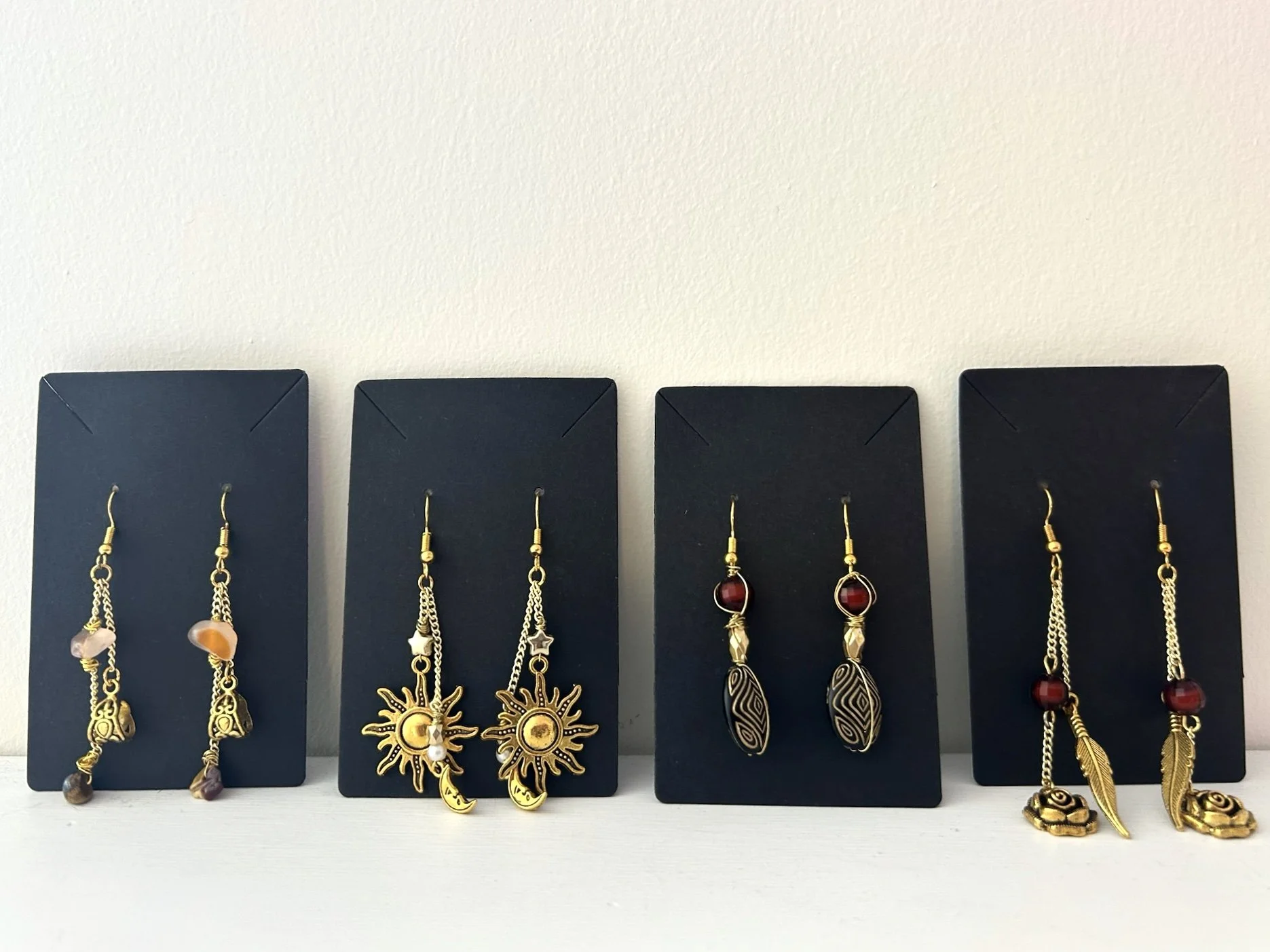 Four pairs of dangling earrings on black display cards, featuring various designs with beads, charms, and intricate patterns.