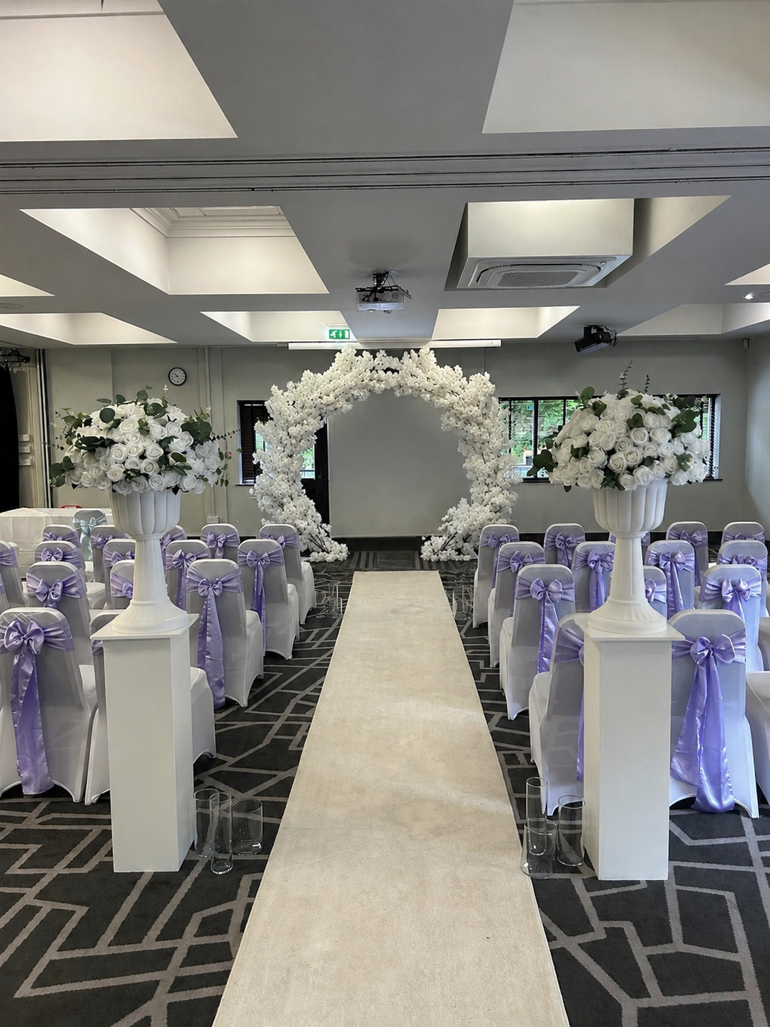 Wedding ceremony setup with an aisle runner, floral arch at the front, and white chairs with purple bows.