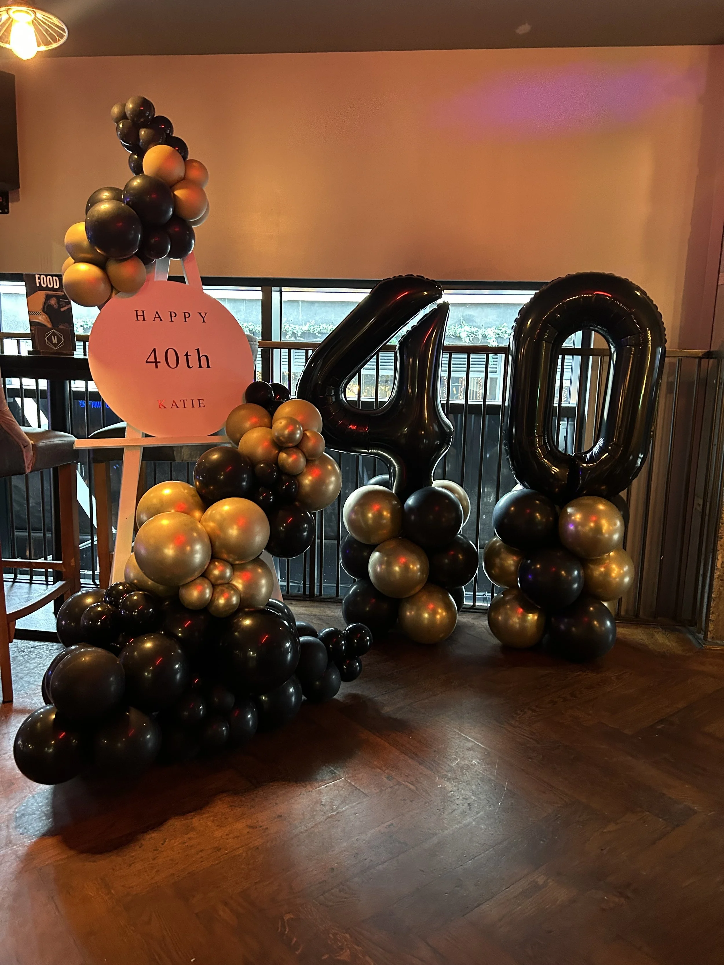 Black, gold, and silver balloons forming the number '40' with a pink sign that reads 'Happy 40th Katie' and balloon arrangements at a birthday celebration.