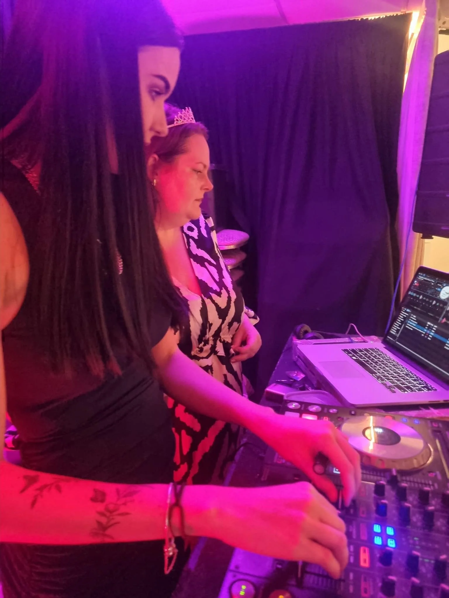 Two women adjusting a DJ mixer and laptop at a party with purple lighting, one wearing black and the other wearing a patterned dress with a tiara.
