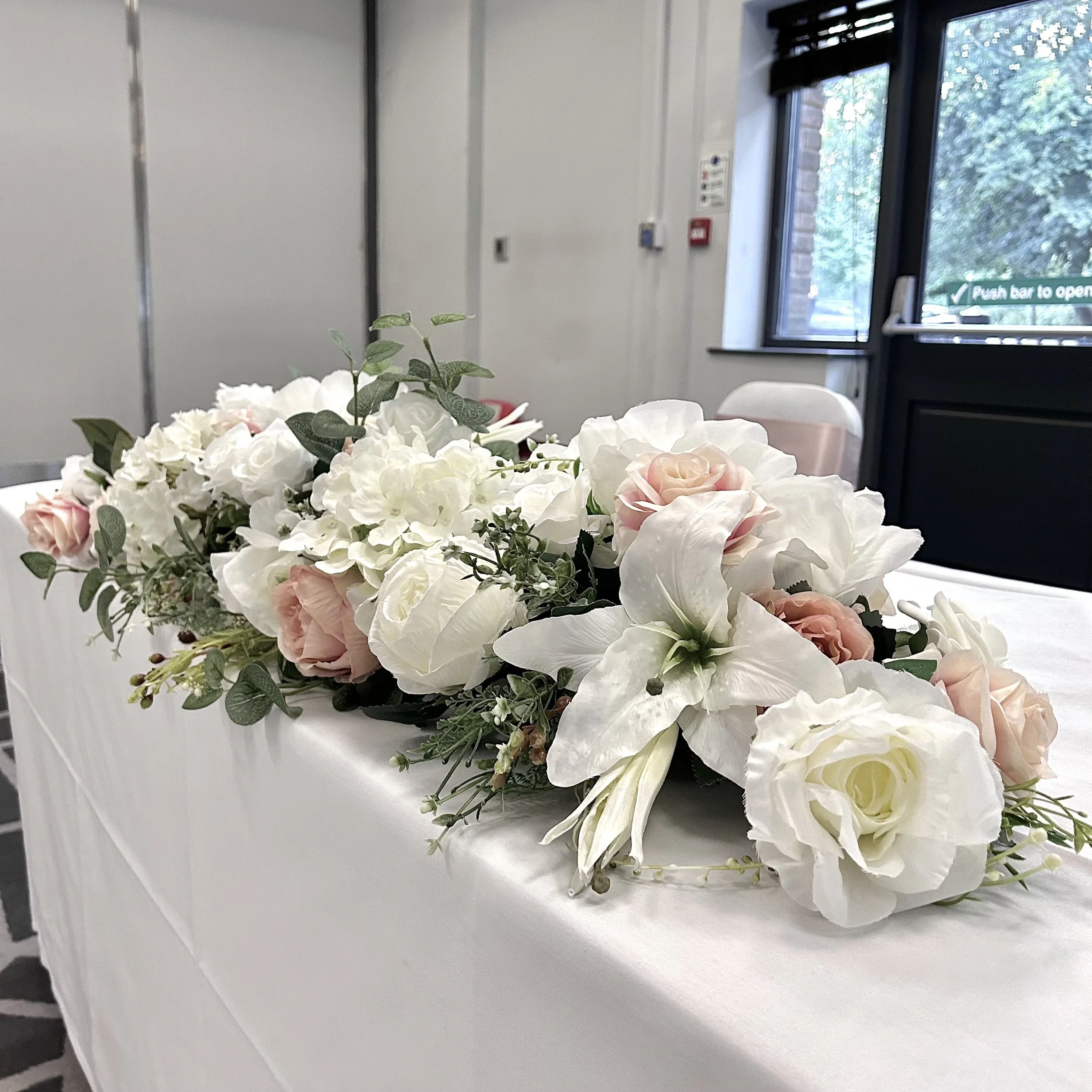 Close-up of a bouquet of white and light pink flowers with green leaves on a white table, inside a room with large windows and a door sign that says 'Push bar to open'.