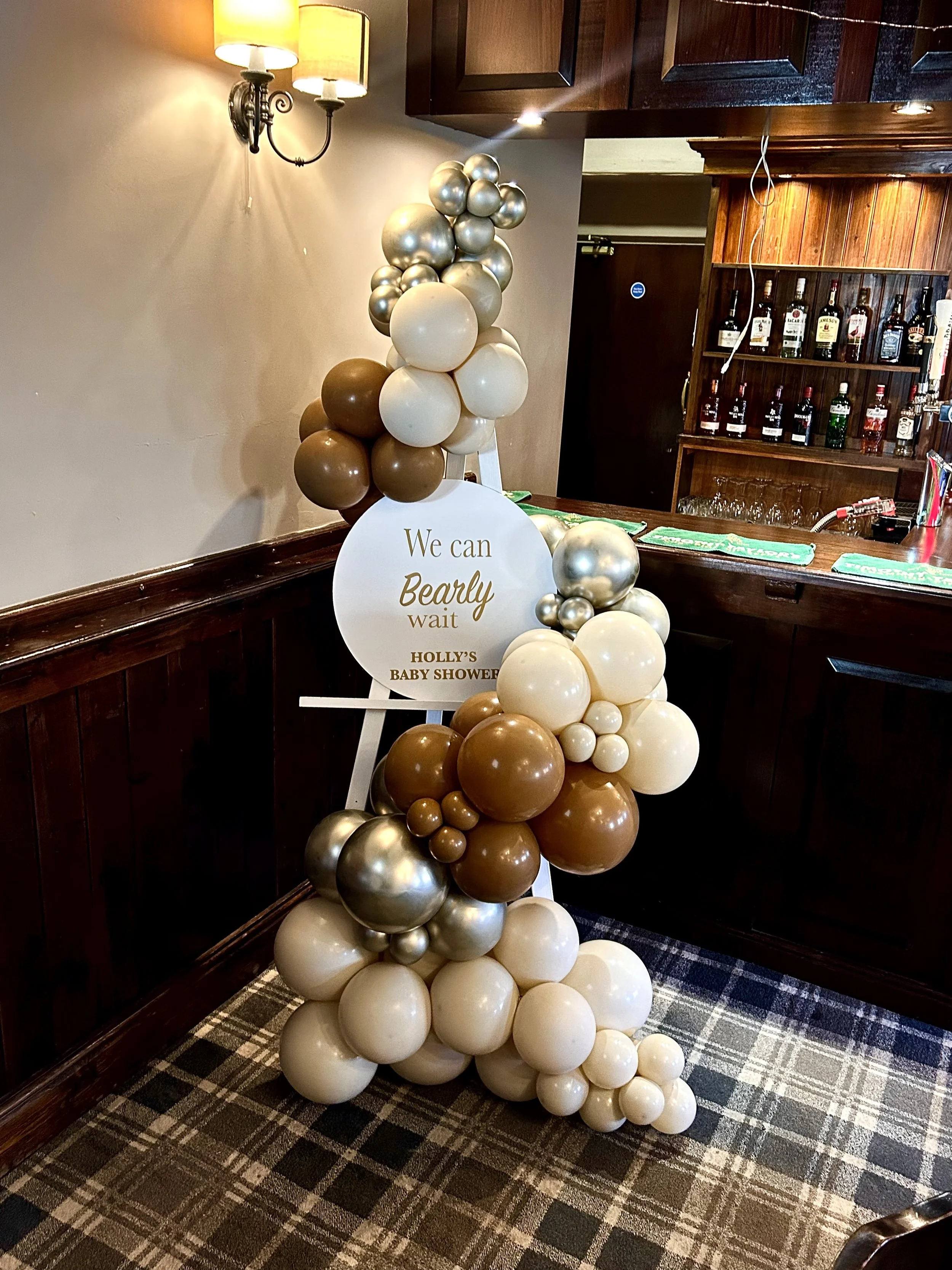 Balloon display with shades of white, cream, brown, and metallic silver, arranged with a sign that reads 'We can Bearly wait, Holly's Baby Shower' in a room with a bar in the background.