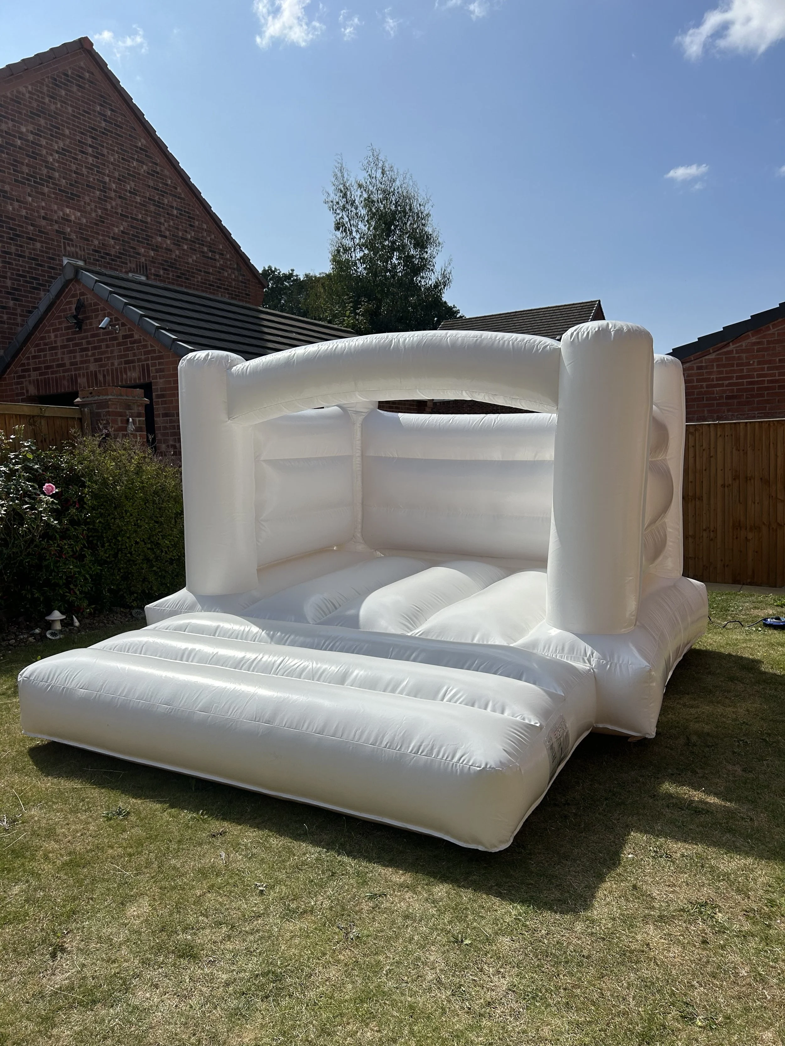 A white inflatable bounce house set up in a backyard with grass, surrounded by a wooden fence, with brick houses and a blue sky in the background.
