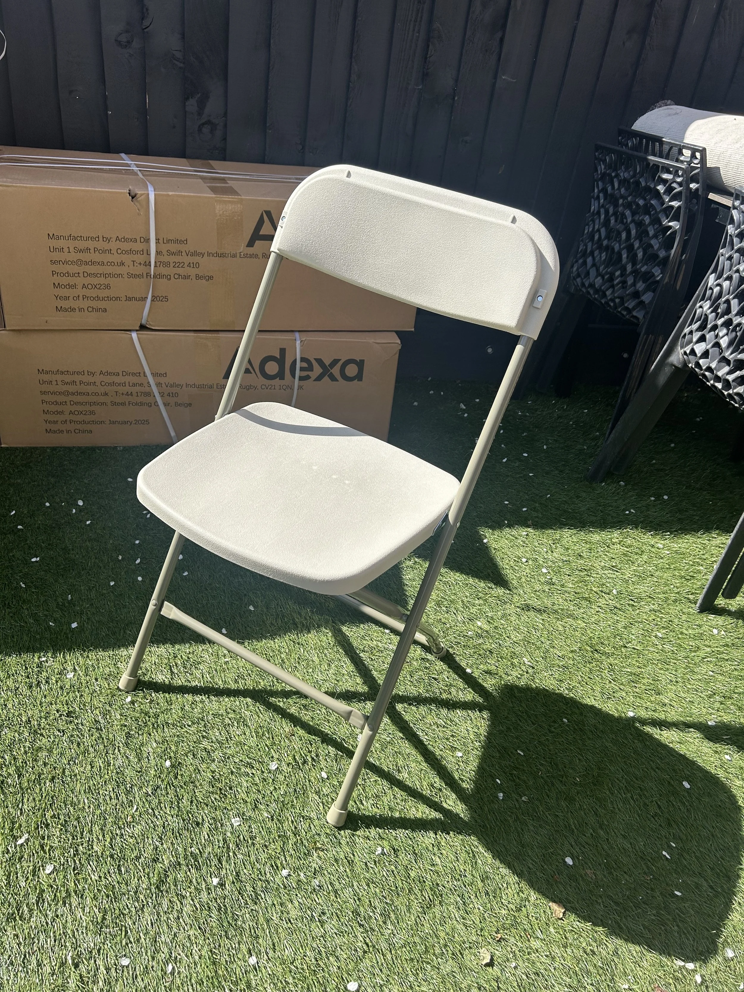 A beige folding chair on a grassy surface, with cardboard boxes and black chairs in the background.