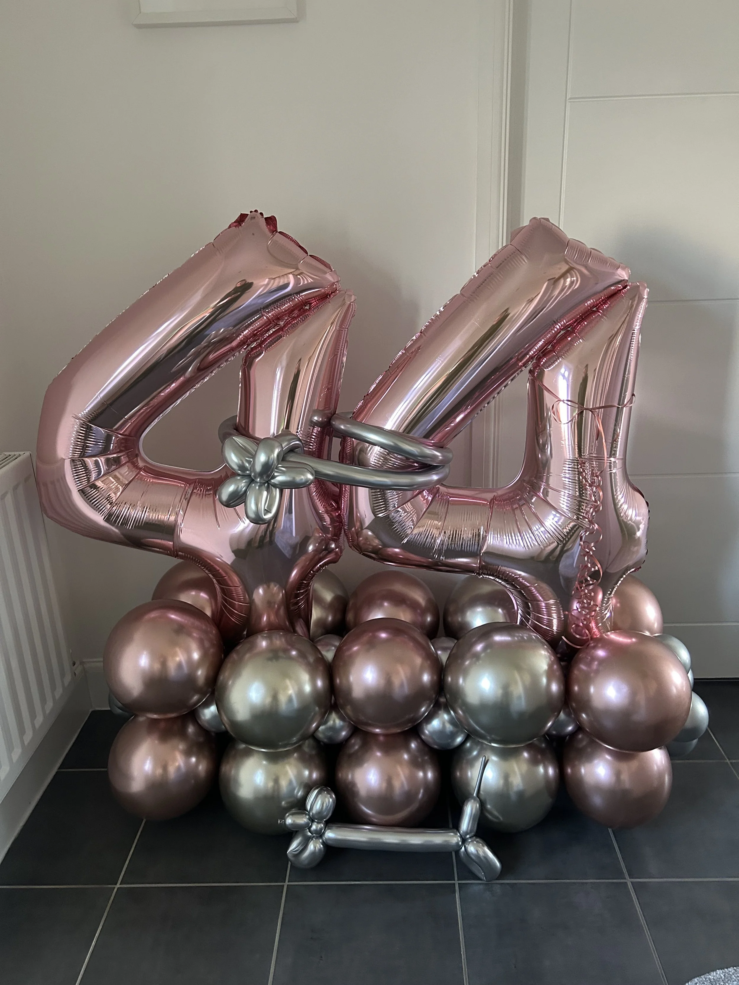 A balloon arrangement featuring large pink metallic number balloons '4' and '4', surrounded by smaller pink and silver metallic balloons, with additional decorative silver and pink balloons on the ground, arranged in front of a plain white wall.