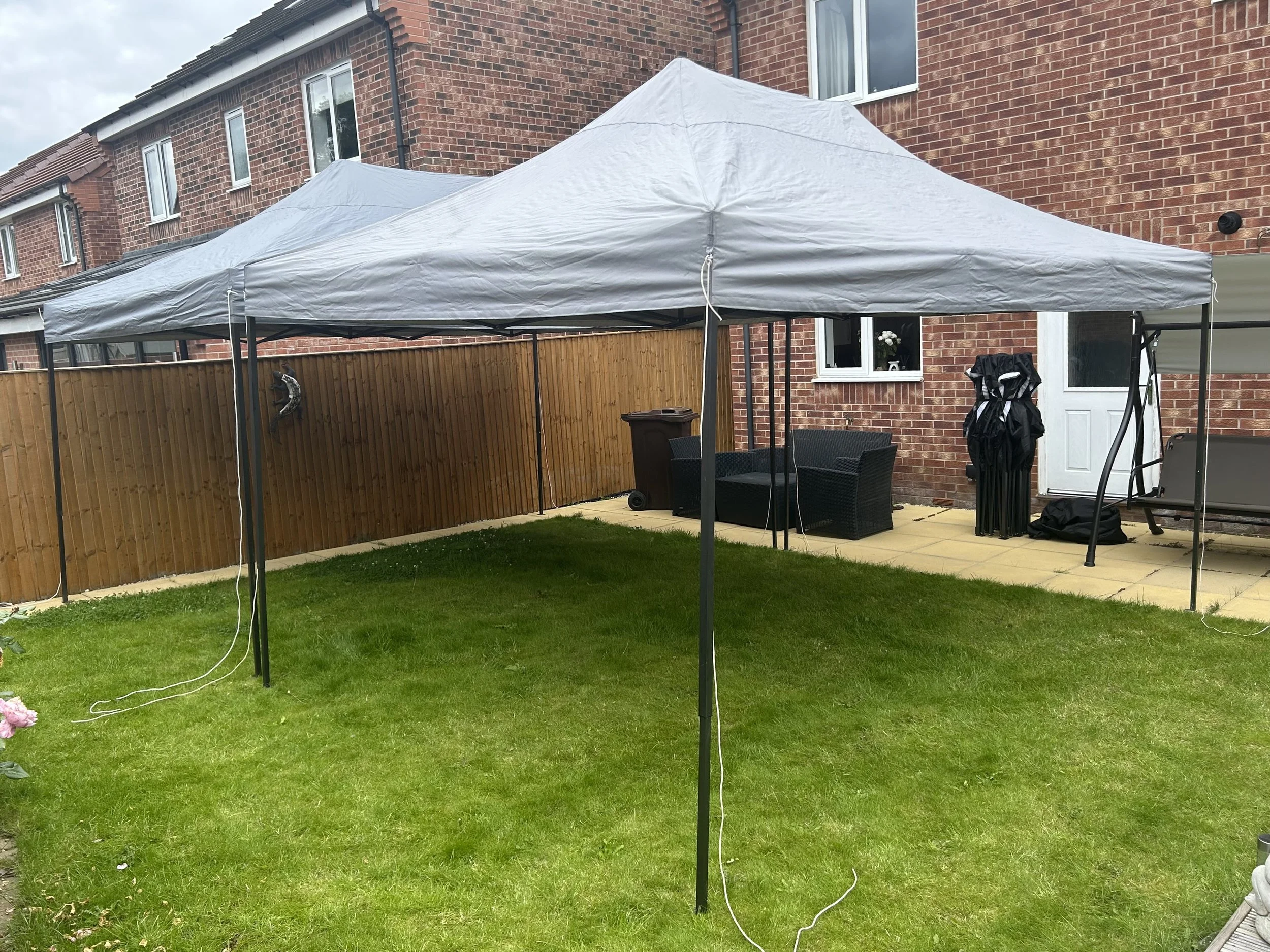 Backyard with a gray canopy tent, green grass, a wooden fence, patio furniture, and a brick house in the background.