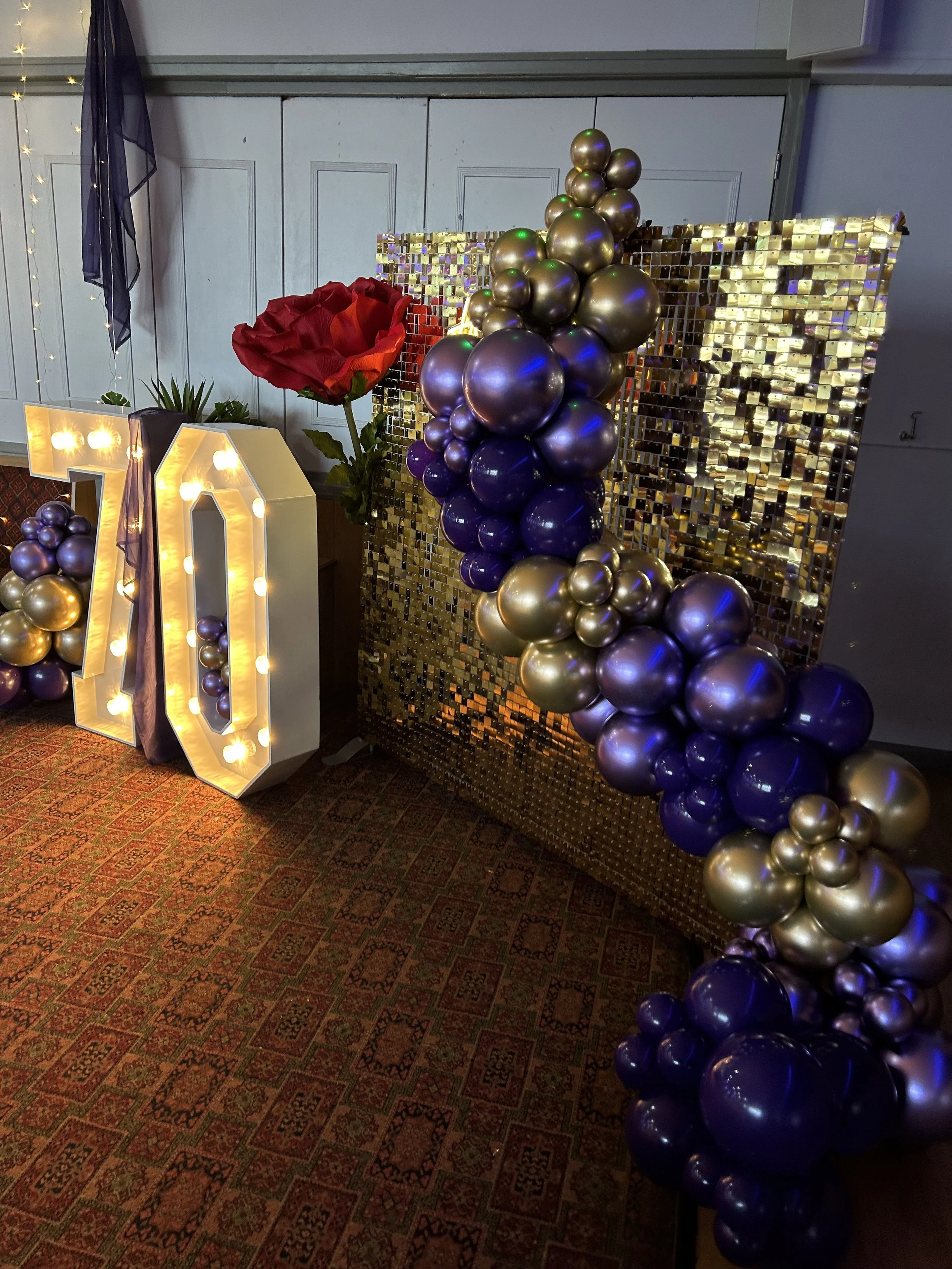 Decorative setup with illuminated '70' sign, purple and gold balloons, a large artificial red flower, a gold mosaic backdrop, and a potted plant in the background.
