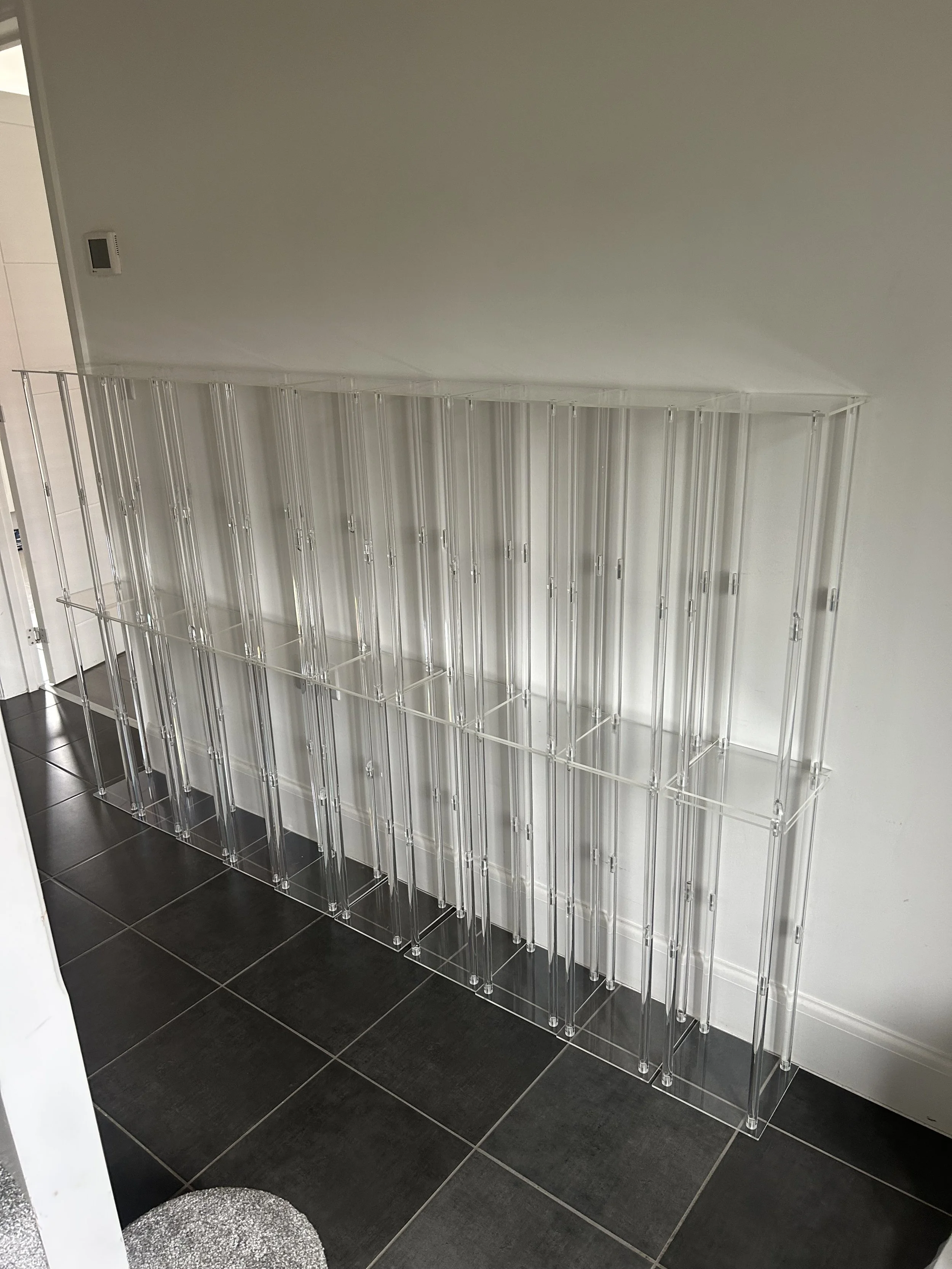 Empty clear acrylic rack with multiple vertical compartments against a white wall on a black tiled floor.