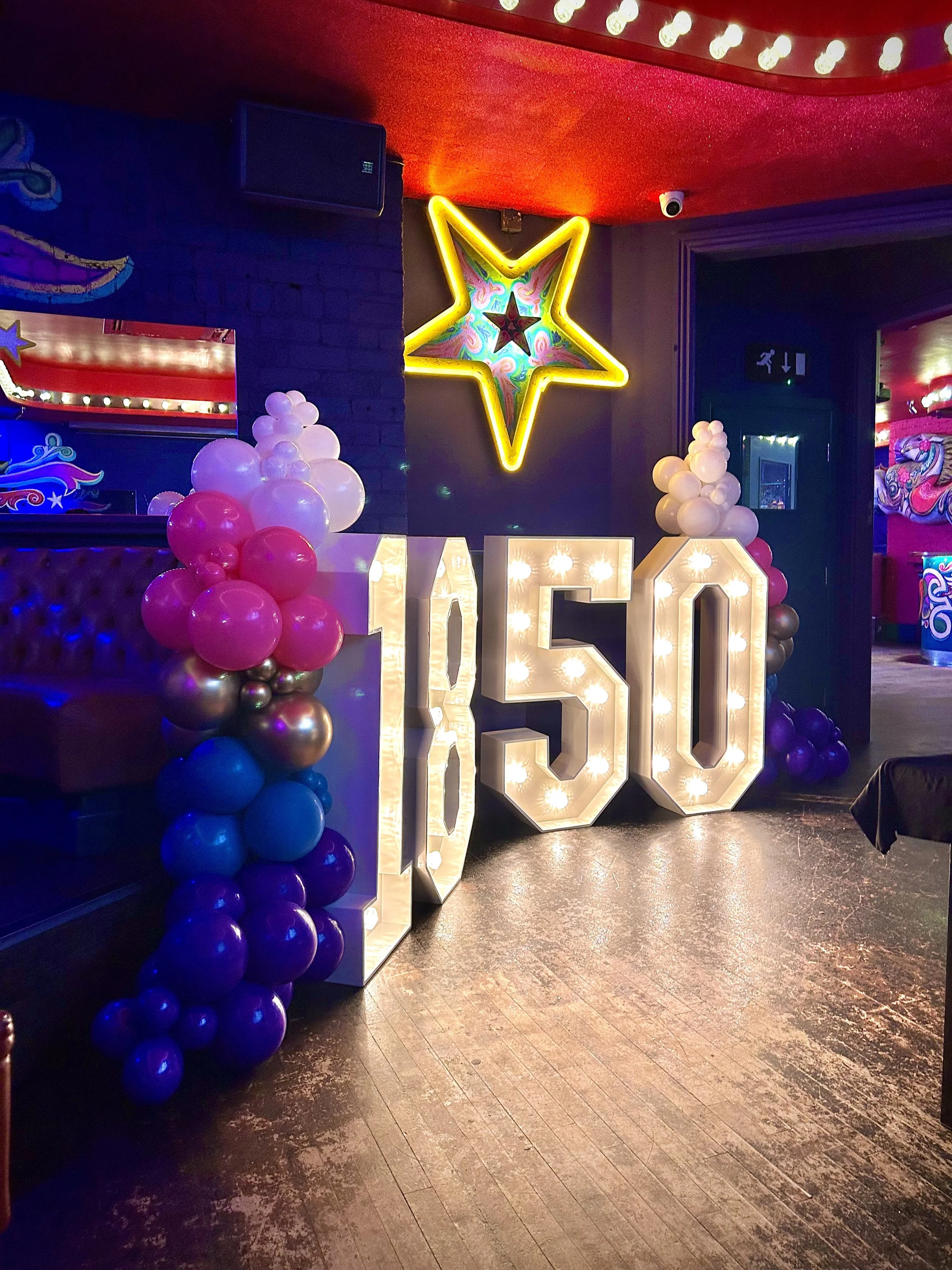 Decorative display celebrating a 50th birthday with large illuminated '50' letters and balloon clusters in pink, purple, blue, white, and gold. A neon yellow star with colorful interior accents hangs on a dark wall in the background.