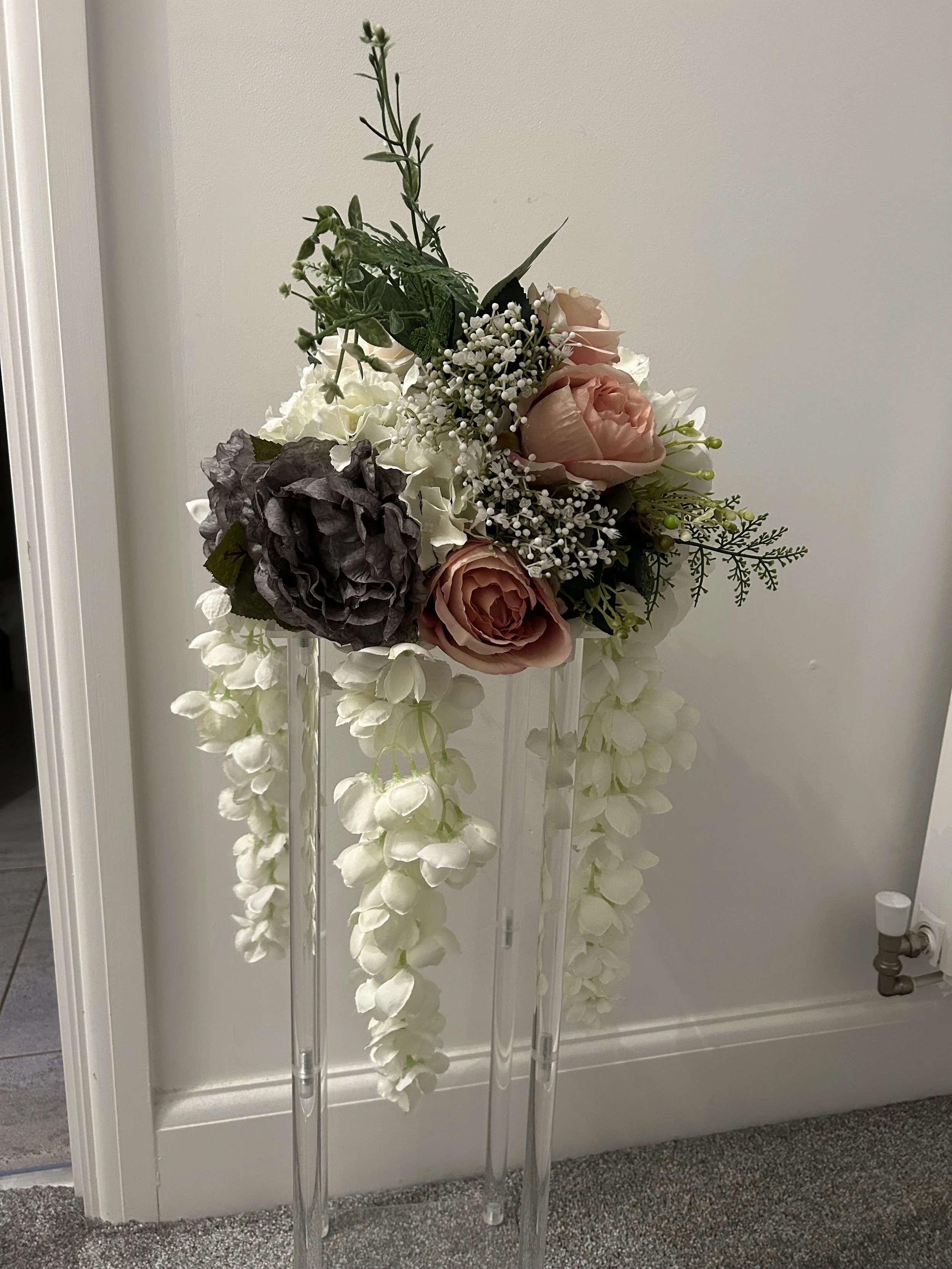 A floral arrangement on clear acrylic stands with pink roses, white hydrangeas, a dark purple peony, and various green foliage.