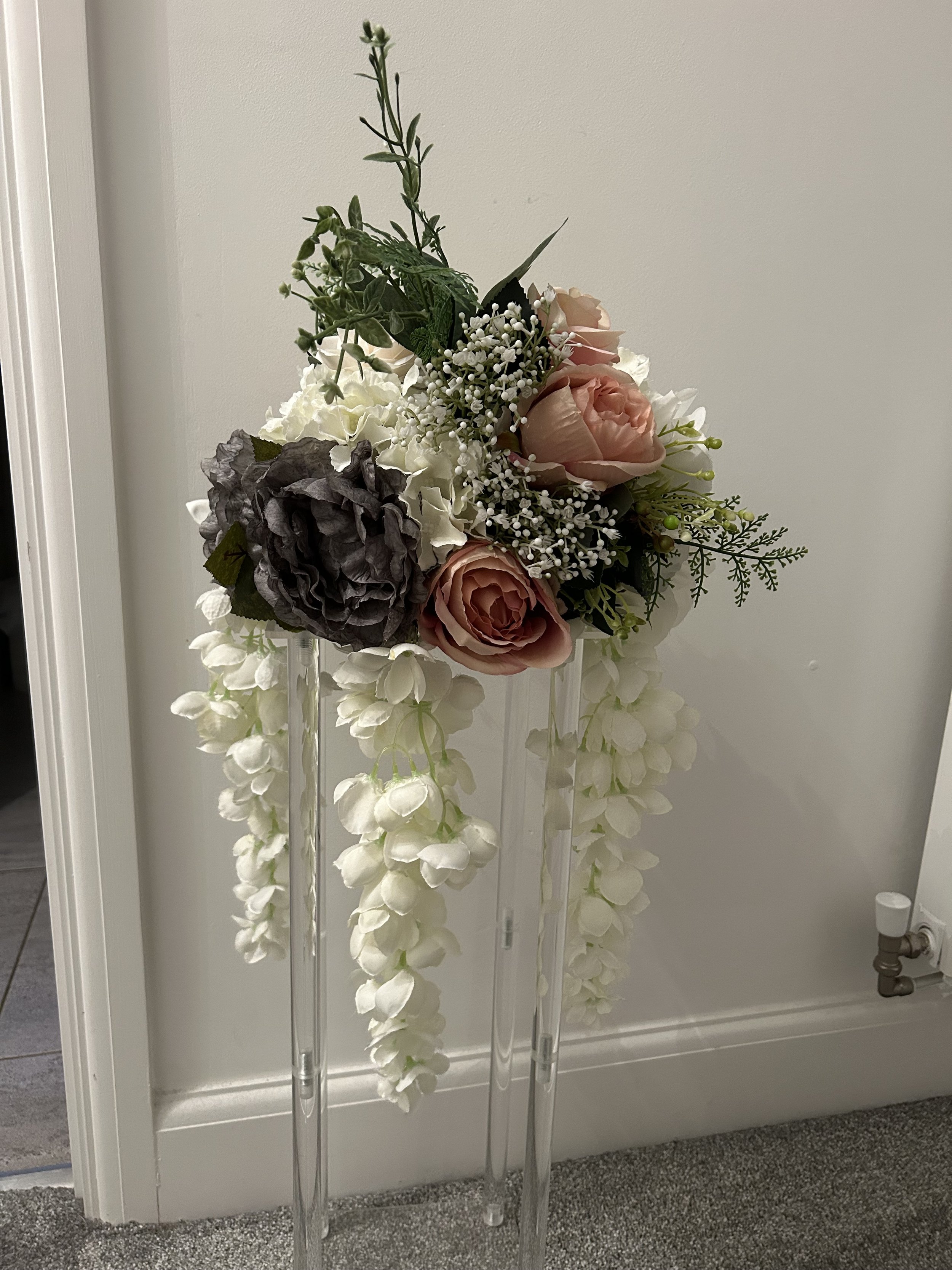 A floral arrangement on tall clear acrylic stands featuring pink roses, white hydrangeas, grayish-purple flowers, small white filler flowers, and various green foliage, placed against a plain white wall near a doorway.