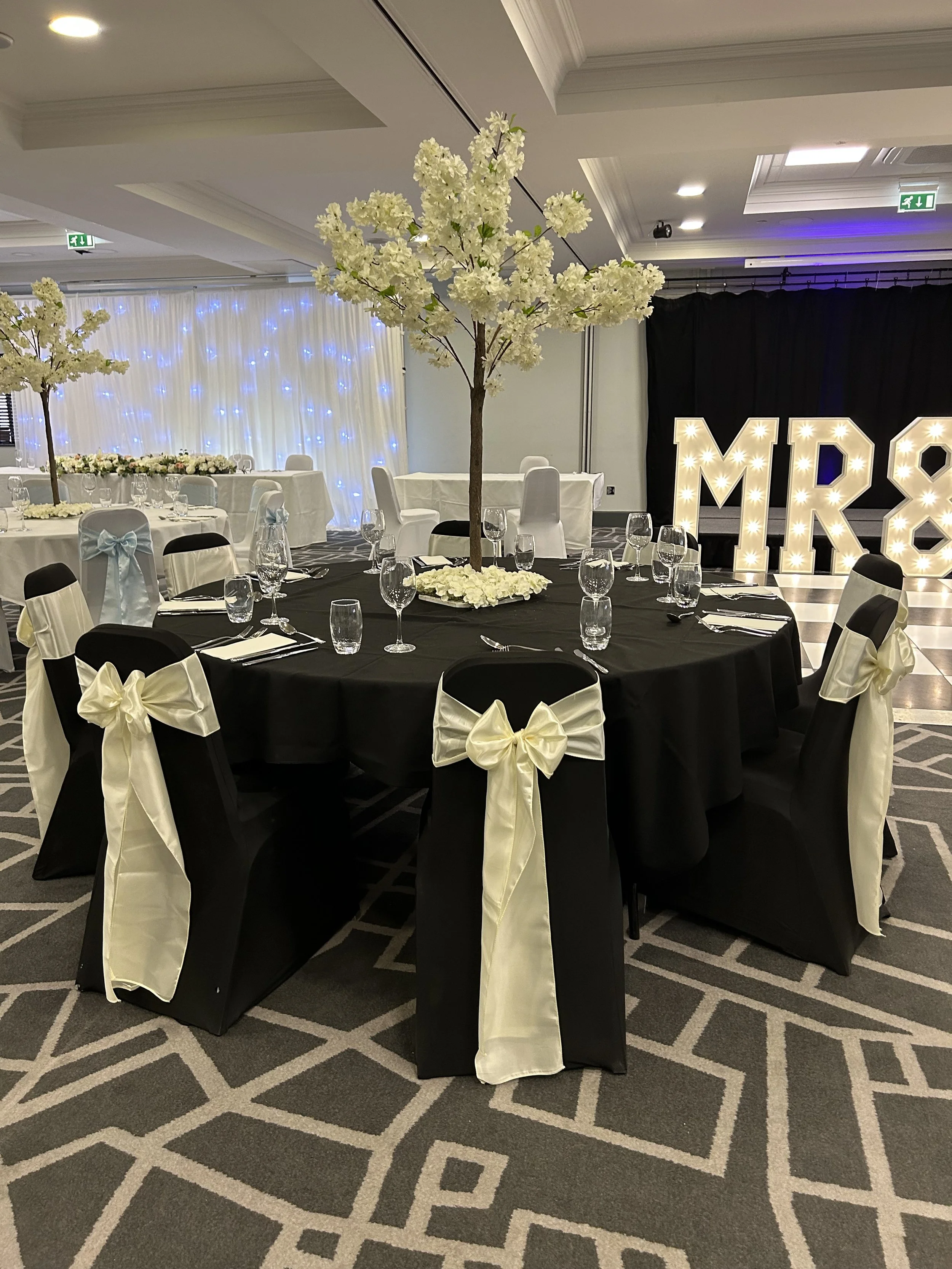 A banquet hall decorated for a wedding reception with white and black chairs, floral centerpieces, and illuminated letter signs that spell 'MR & MRS'.