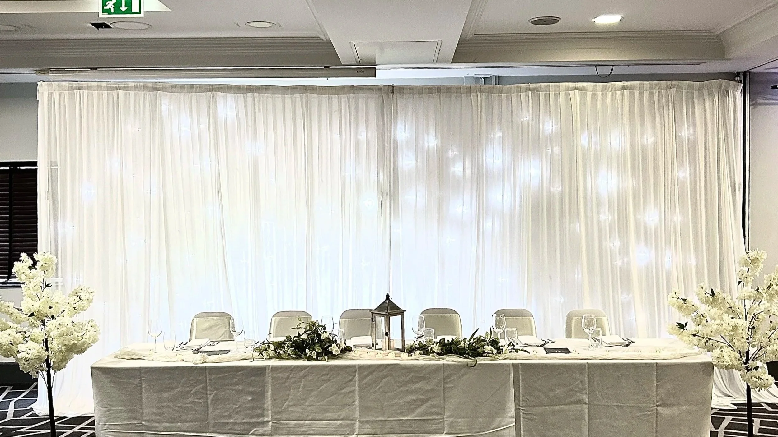 A wedding or event reception table setup with white tablecloth, floral centerpiece, lantern, wine glasses, plates, and napkins, in front of a white curtain backdrop with lights.