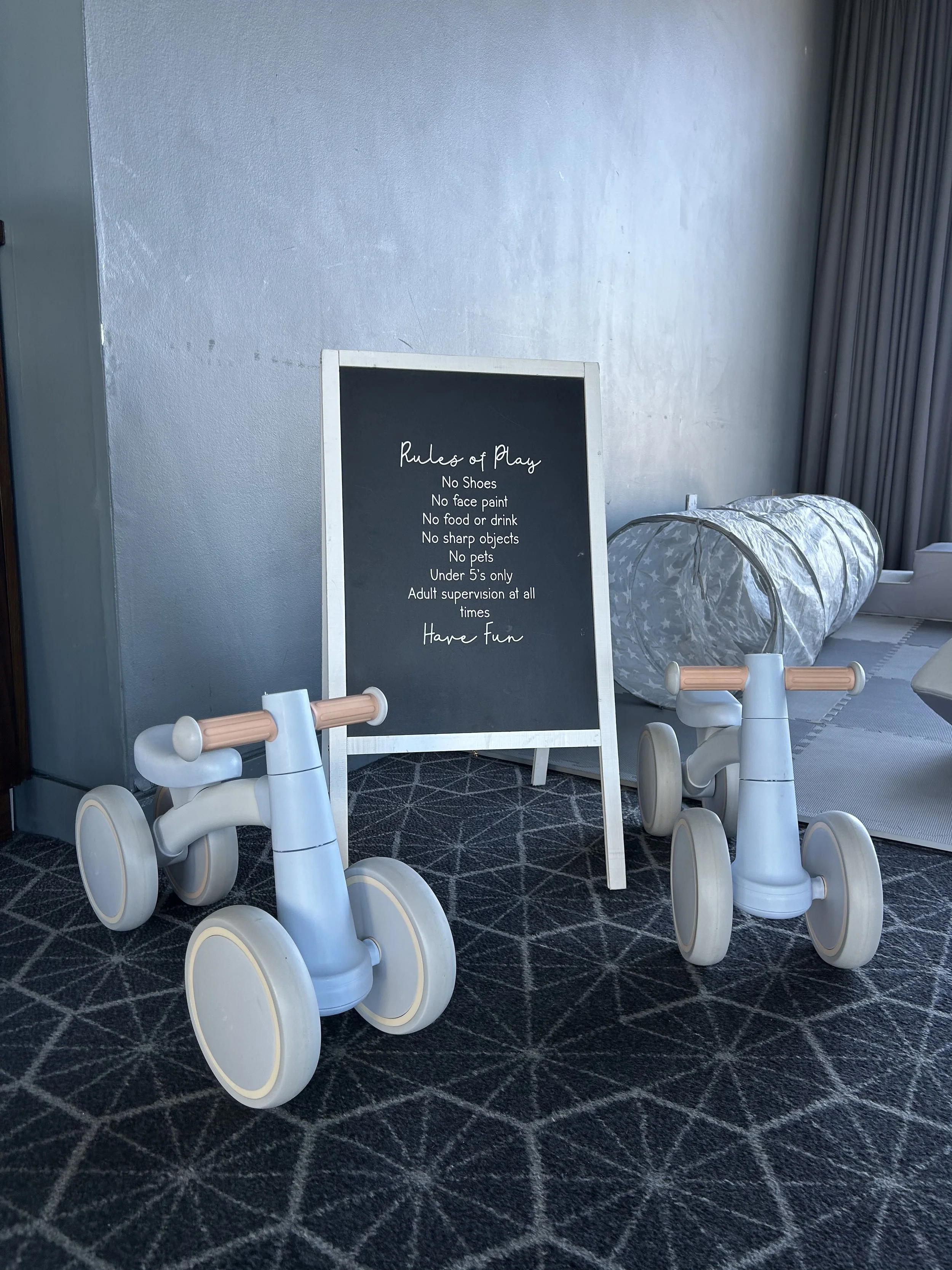 Indoor play area with two small ride-on toys, a blackboard with rules for play, and a soft tunnel in the background. Soft play hire