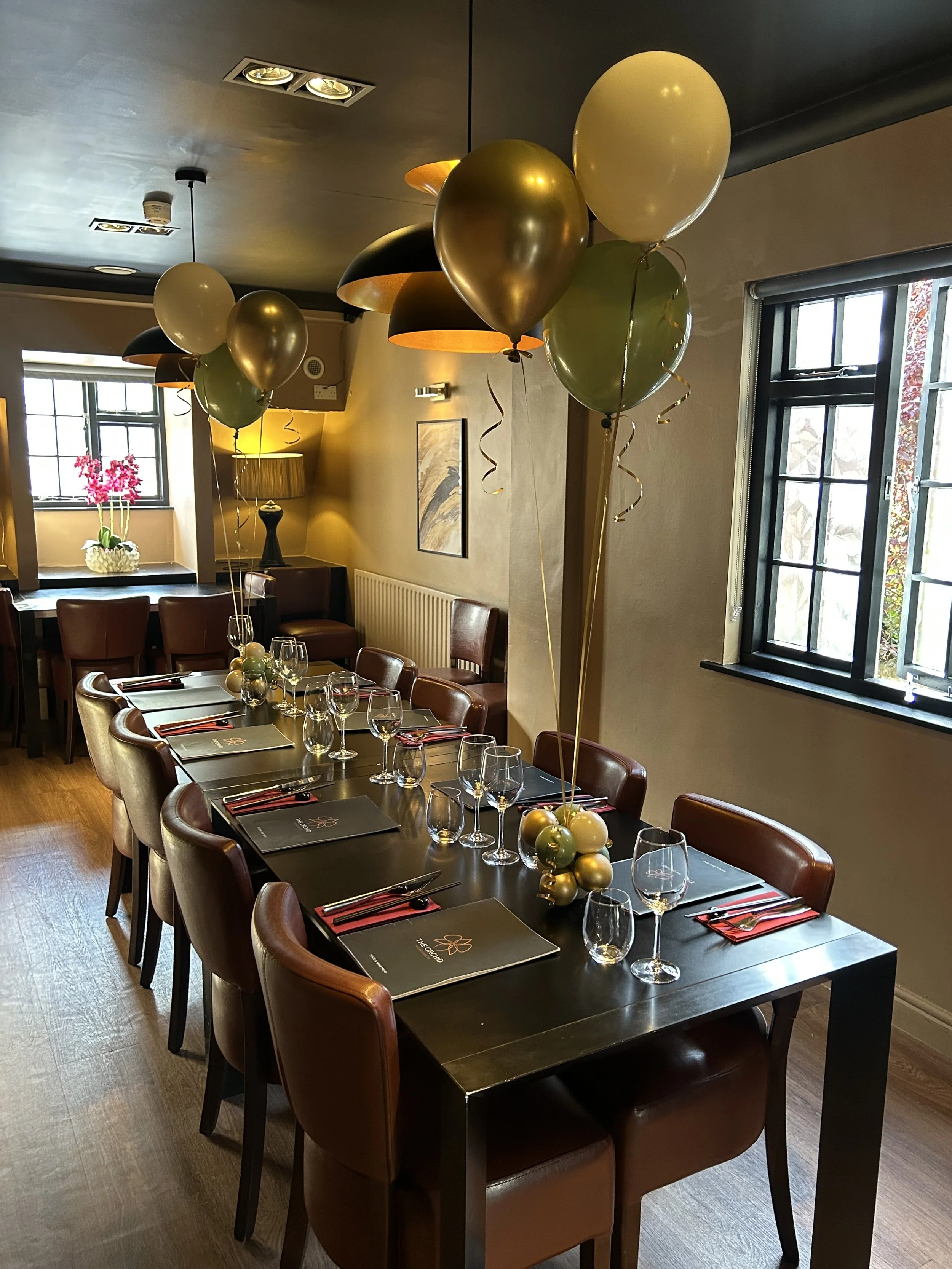 Party table set with wine glasses, black menus, and red napkins, decorated with floating balloons in black, gold, white, and green.