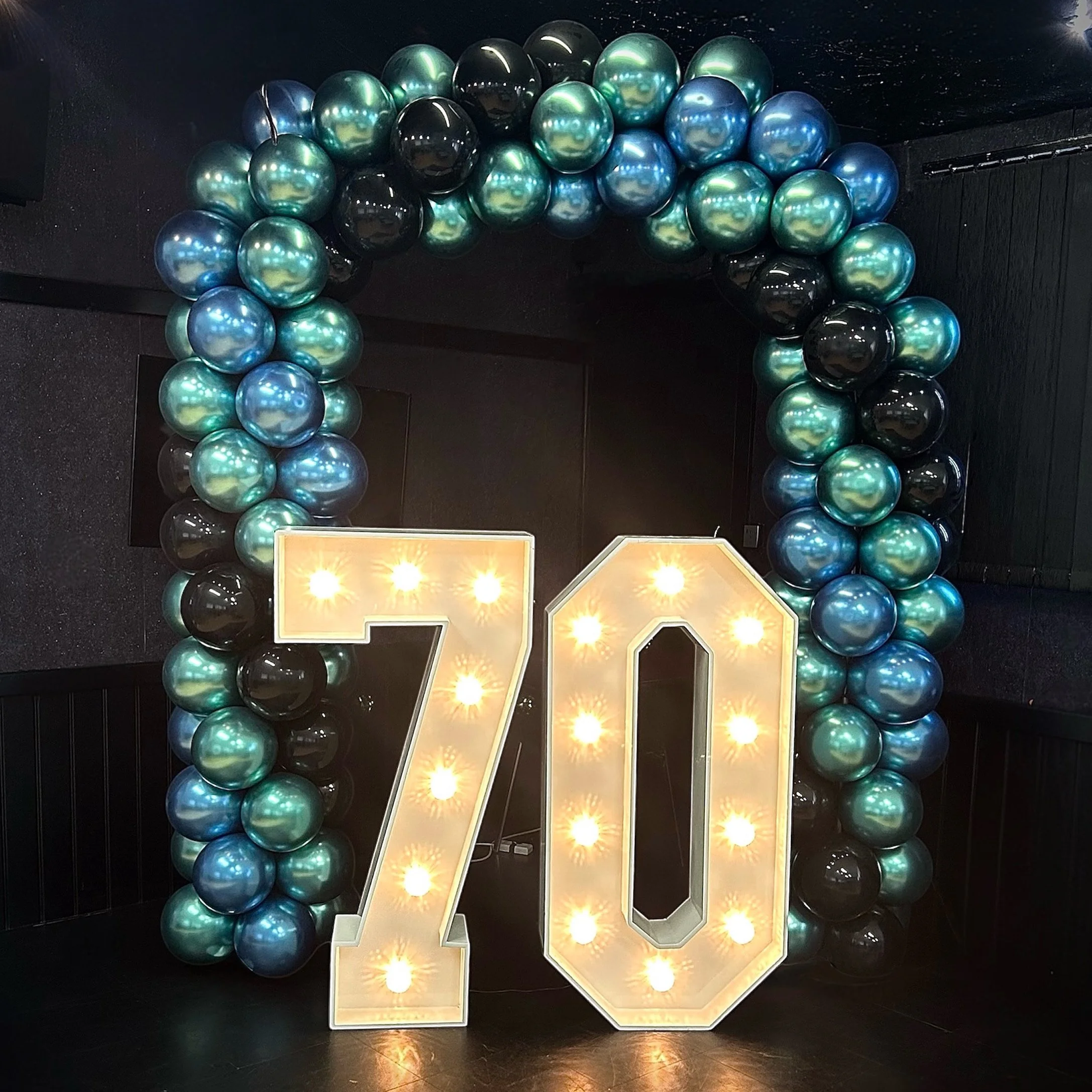 Decorative birthday display with a large illuminated "70" and a balloon arch in shades of teal, black, and blue.