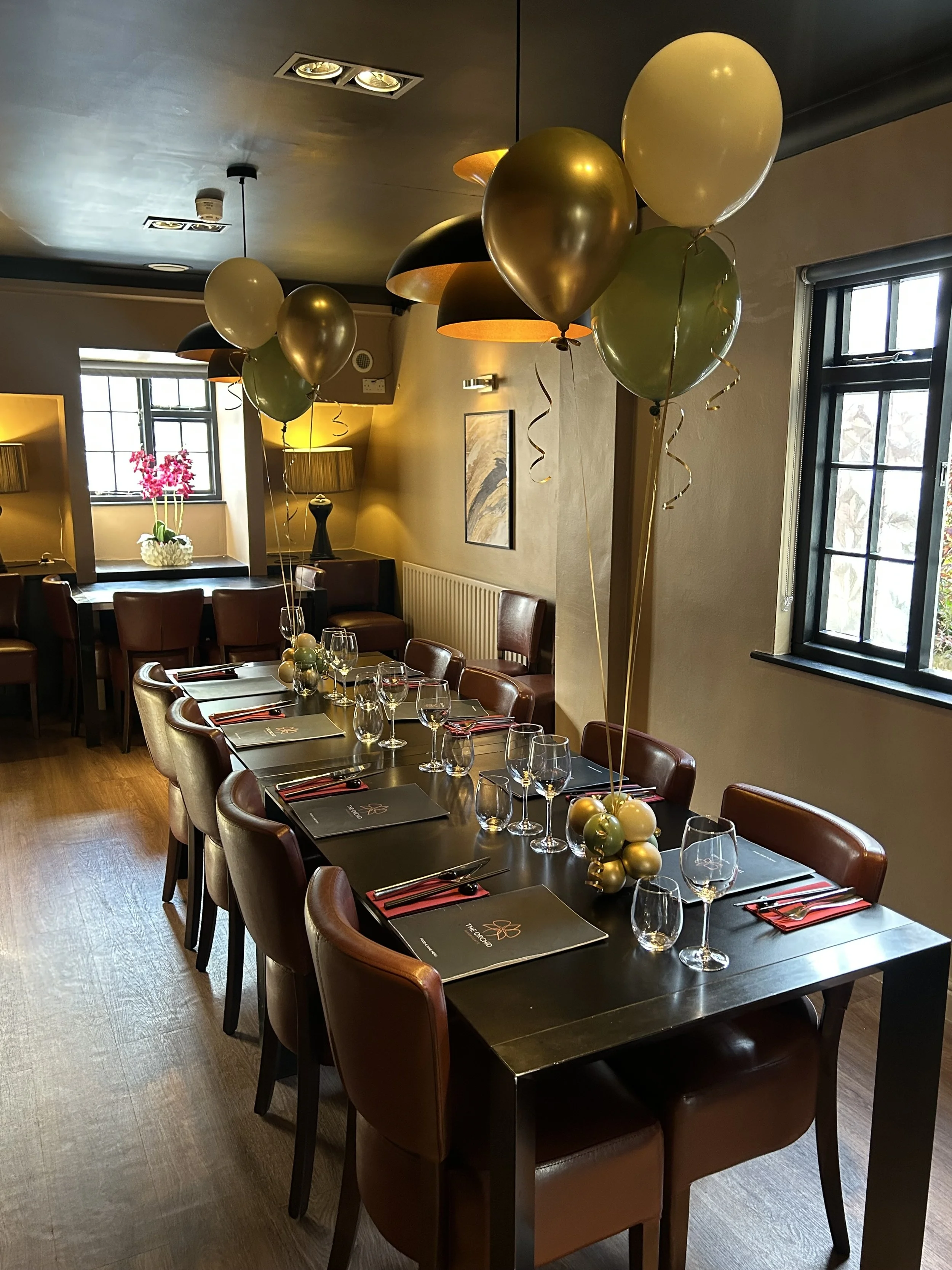 Decorated dining table with black placemats, wine glasses, and balloons in gold, green, white, and black in a nicely lit restaurant or dining room.