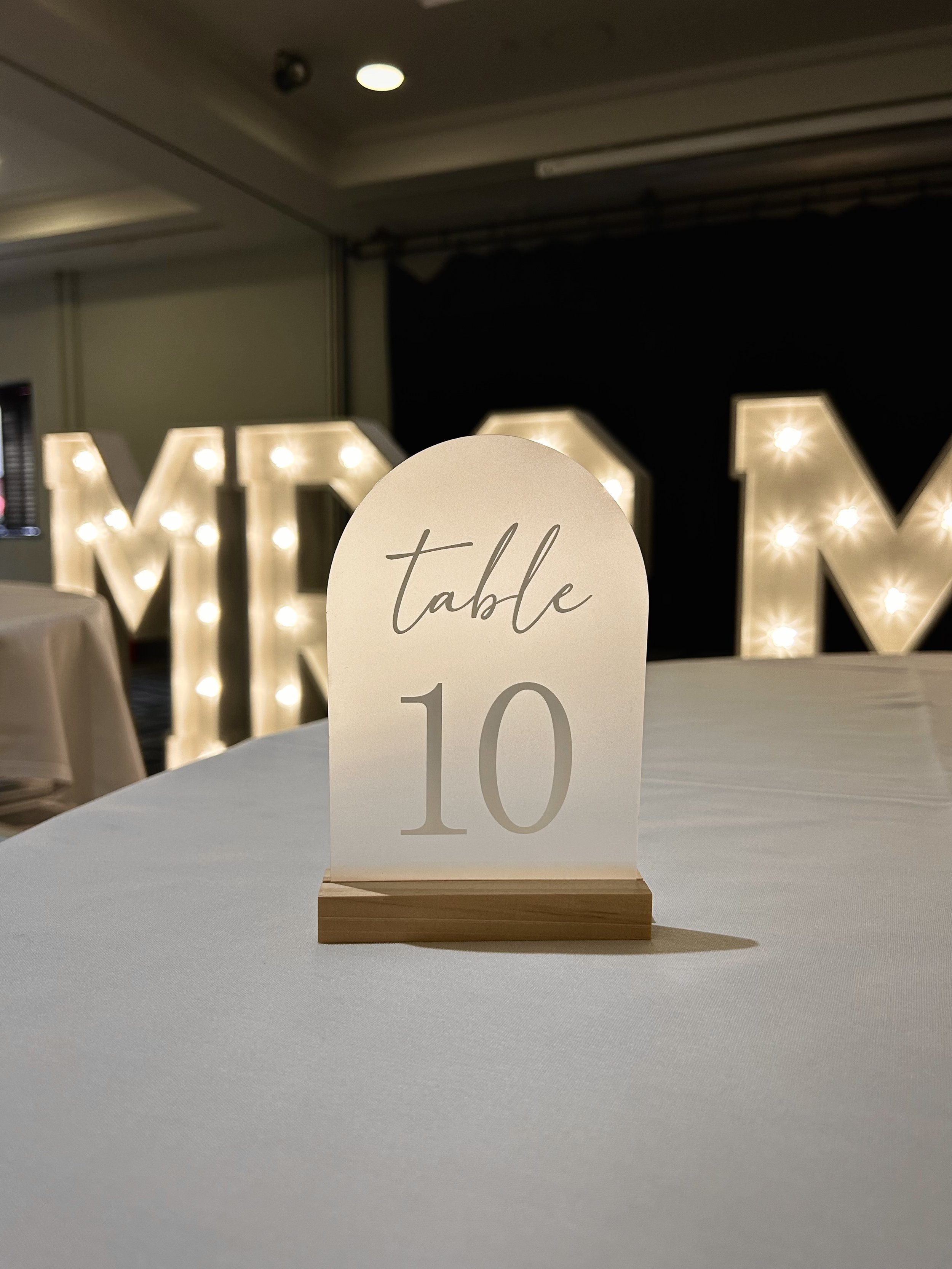 Table number card on a white tablecloth, with the number 10 and the word 'table' written on it, in a banquet or event setting with illuminated decorative letters in the background.