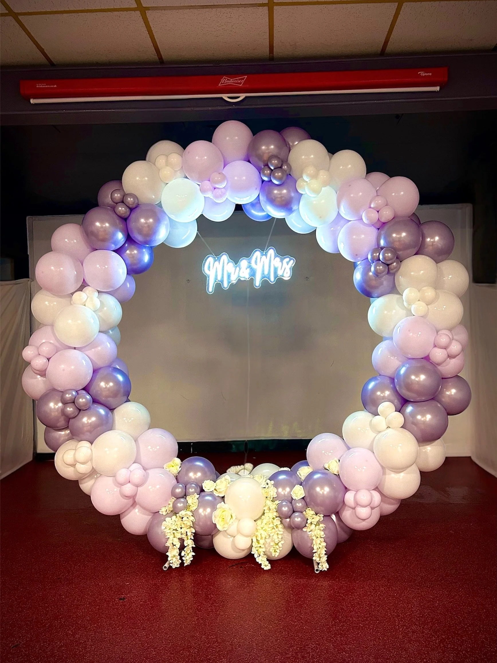 A circular balloon arch in shades of purple, white, and silver, decorated with flowers at the bottom, with a neon sign that reads 'Mrs. & Mr.' in the background.