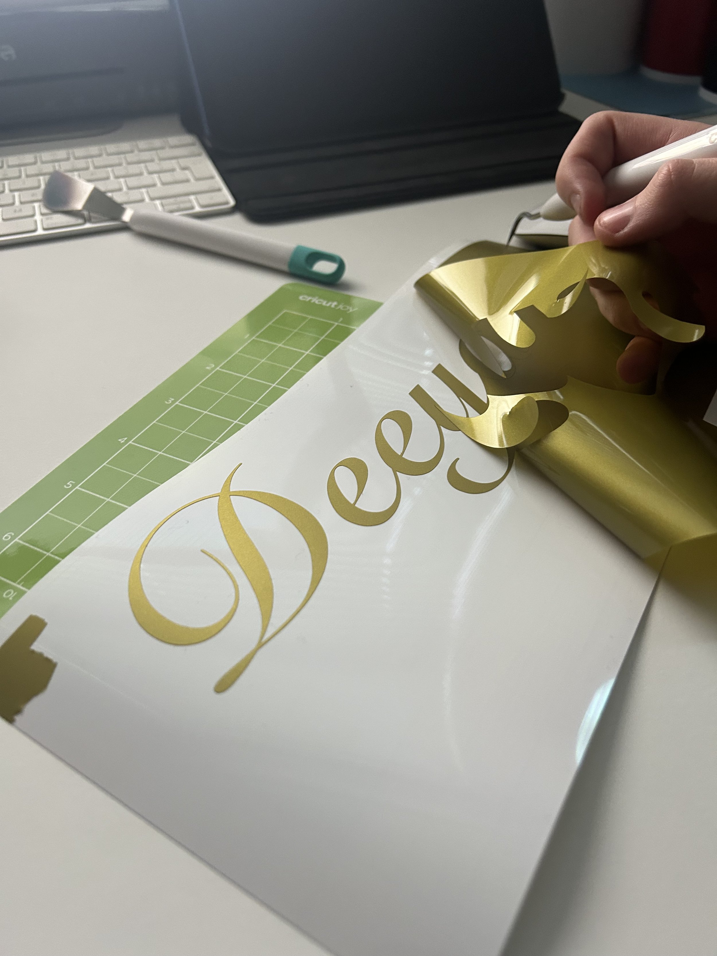 A person is applying gold and white vinyl decals to a white surface for crafting, with a green cutting mat, a white pen, and other crafting tools on the table.