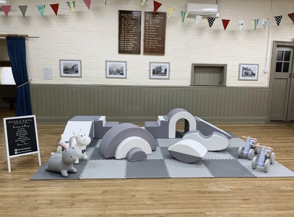 Children's indoor play area with soft play equipment, including animal-shaped seats, arch-shaped structures, and small wooden tricycles, on a checkered gray and white foam mat. The room has white brick walls with framed black-and-white photographs, colorful pennant banners hanging from the ceiling, a blue curtain on the left, and a wooden plaque with names on the wall.