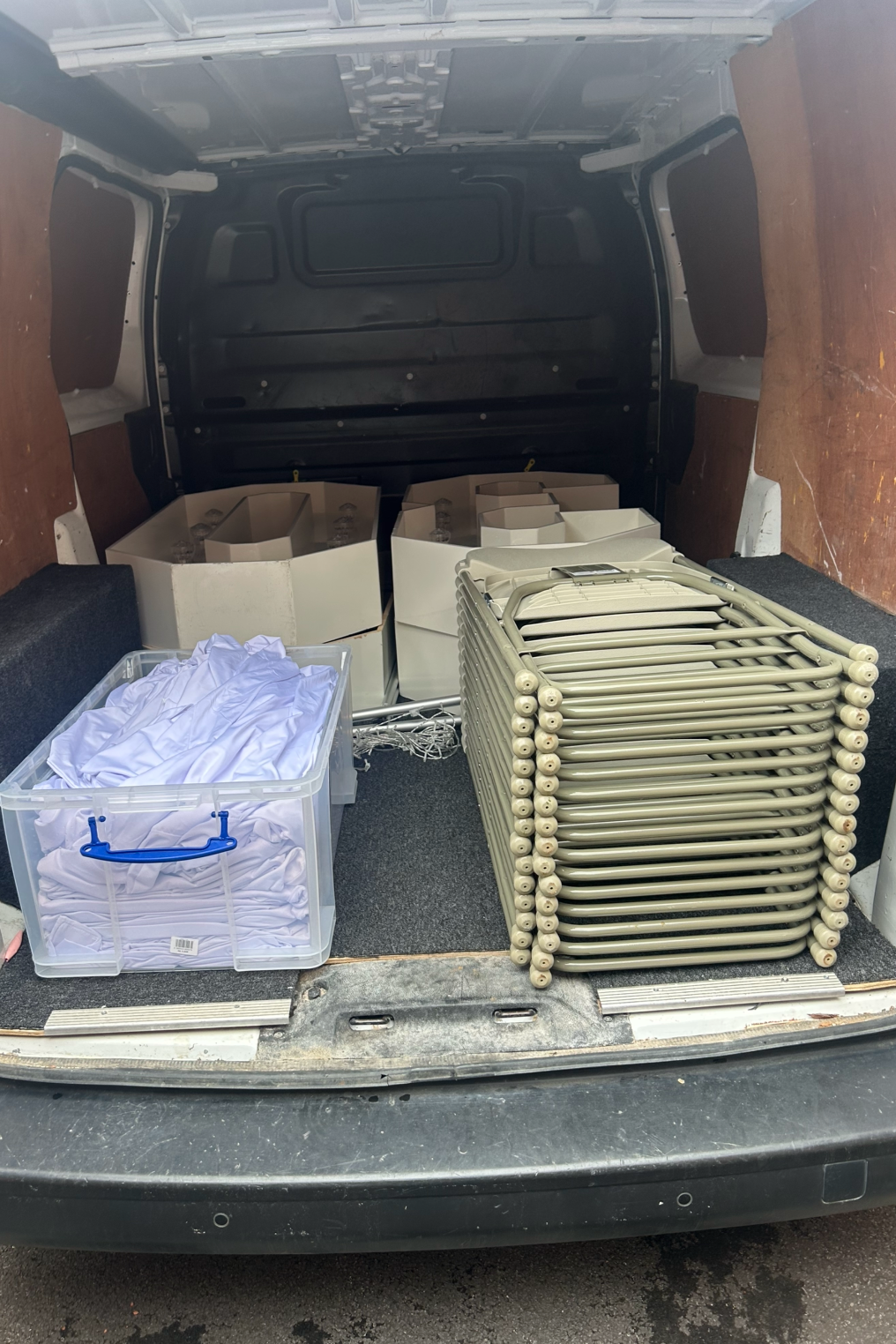 The back of a van loaded with plastic folding chairs, white boxes filled with white fabric, and a plastic bin containing white shirts.