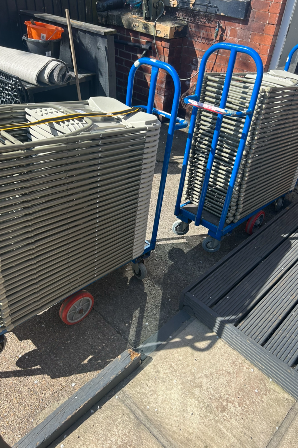 Trolley carts stacked with metal chairs and benches outside on pavement, next to a brick wall and black building entrance, with shovels, containers, and other tools in the background.