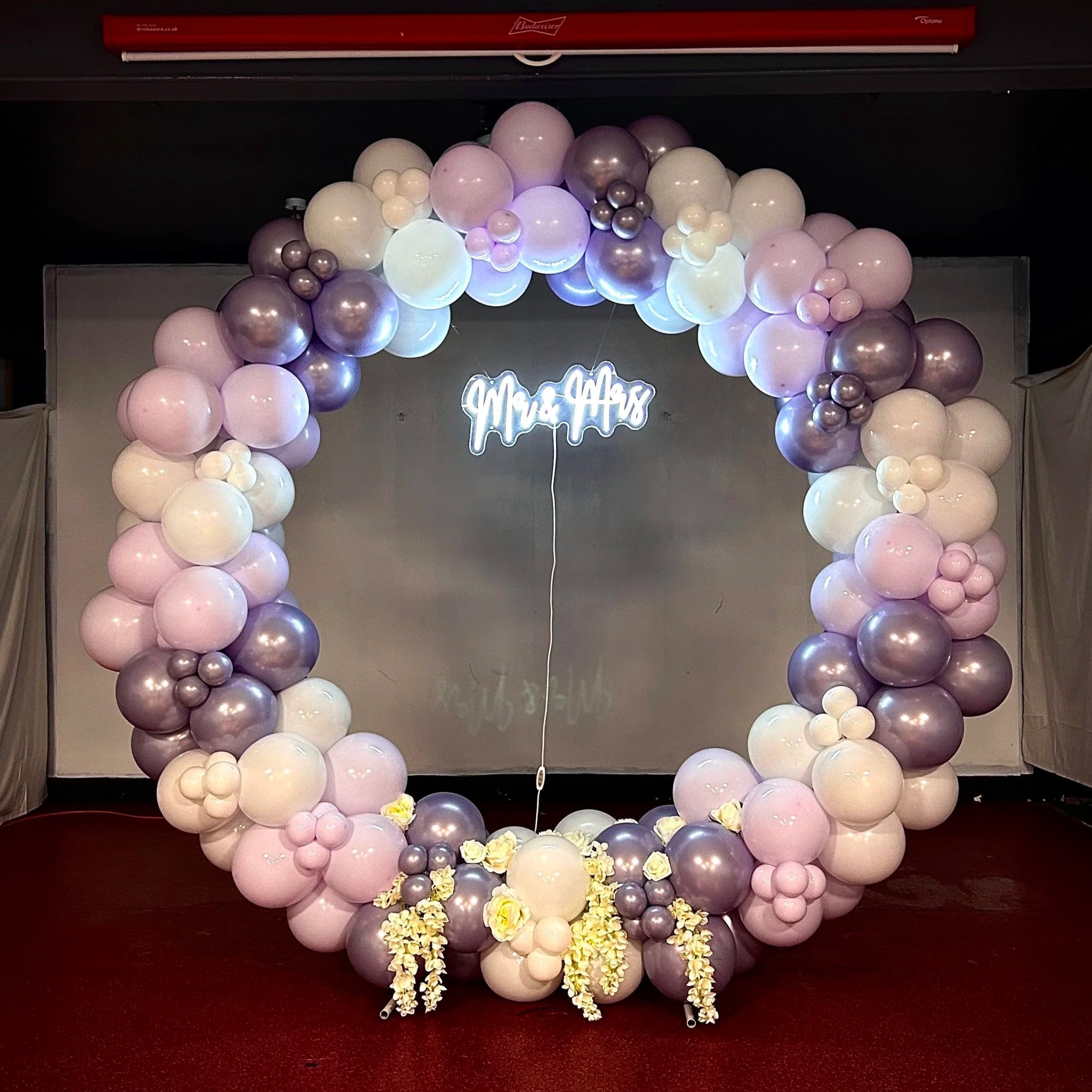 Decorative balloon arch in shades of pink, white, and metallic silver with flowers at the base, set against a dark background with a sign reading 'Mr. & Mrs.' and a red ceiling above.