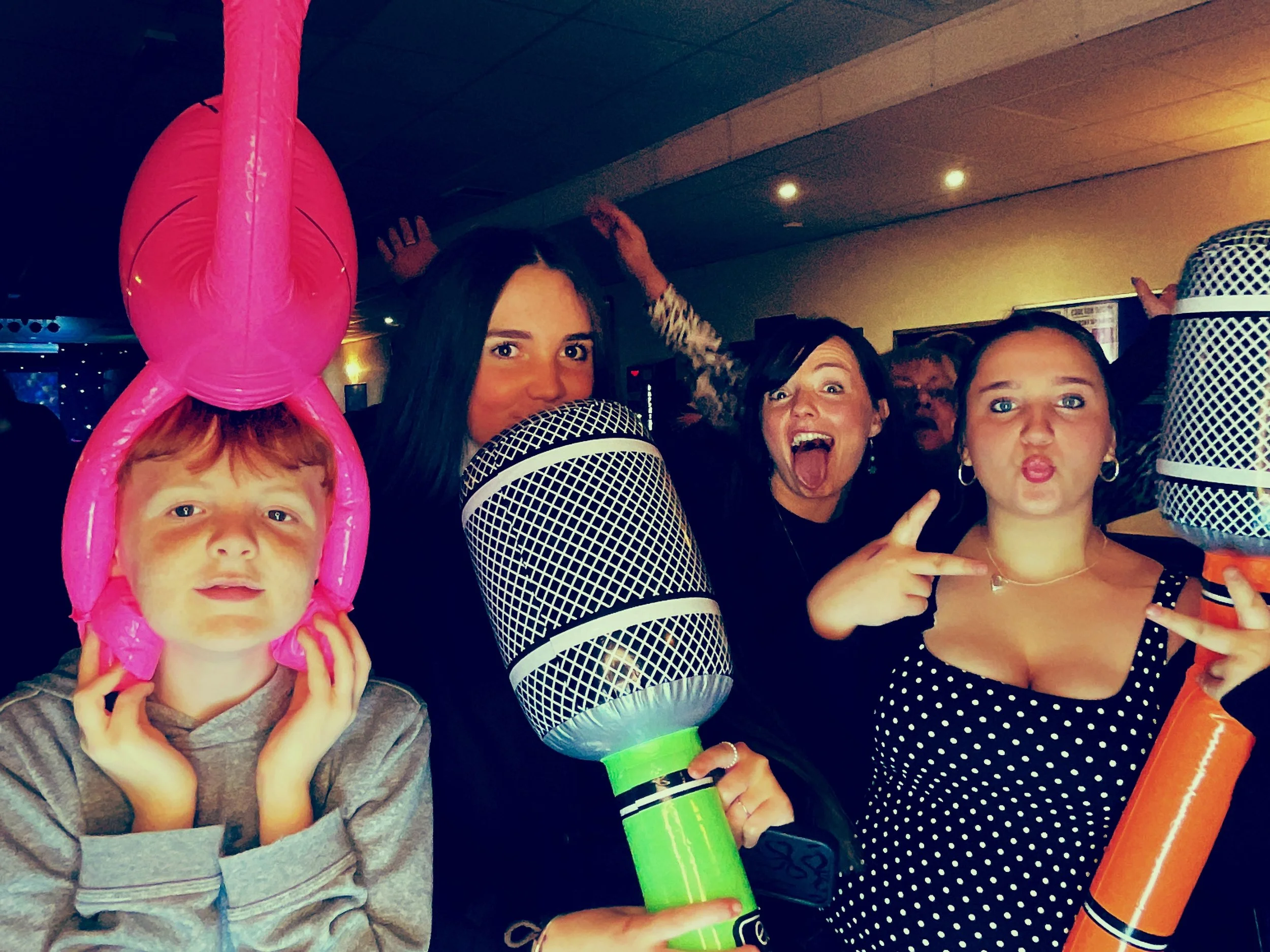 Group of people celebrating at a party with balloons and props, including a young boy with pink balloon hat, and women making playful expressions and gestures.