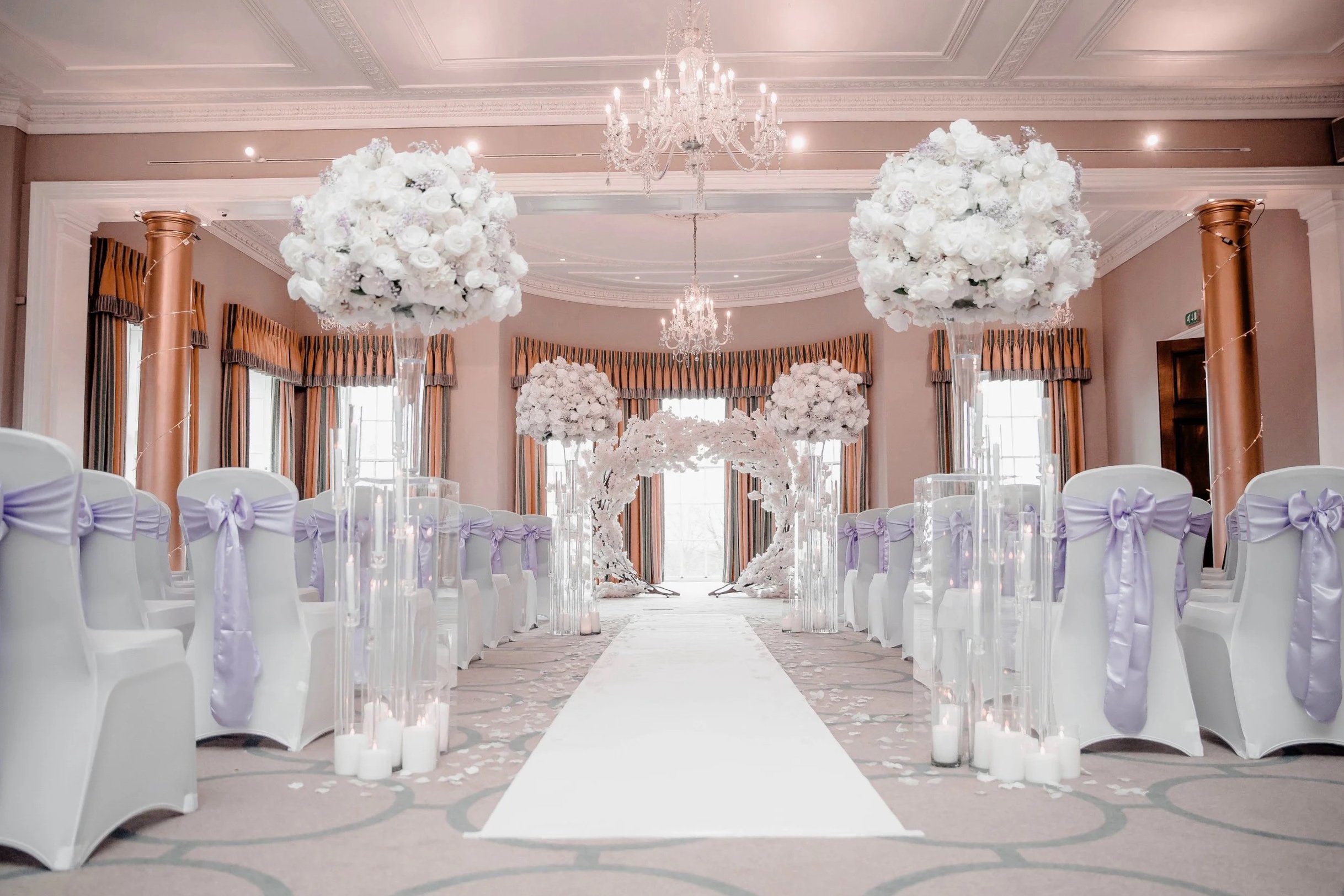 Elegant wedding reception with white chairs adorned with lavender ribbons, floral centerpieces, tall candles, a white aisle runner, chandeliers, and draped windows in a decorated ballroom.