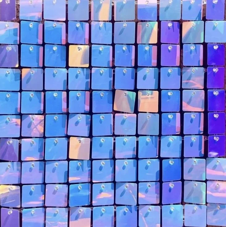 A close-up of many small, square, iridescent tiles arranged in a grid. The tiles reflect shades of blue, purple, pink, and orange, creating a colorful and shiny mosaic pattern.