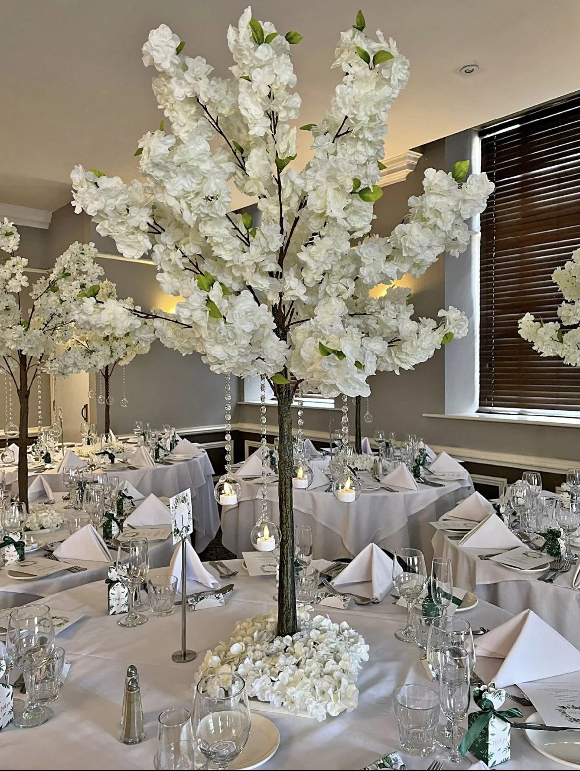 A banquet table decorated with a large white floral centrepiece blossom trees surrounded by white napkins, glassware, and candles in a well-lit room with dark window blinds.