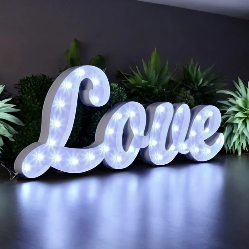 White illuminated decorative sign spelling 'Love' with green potted plants in the background.