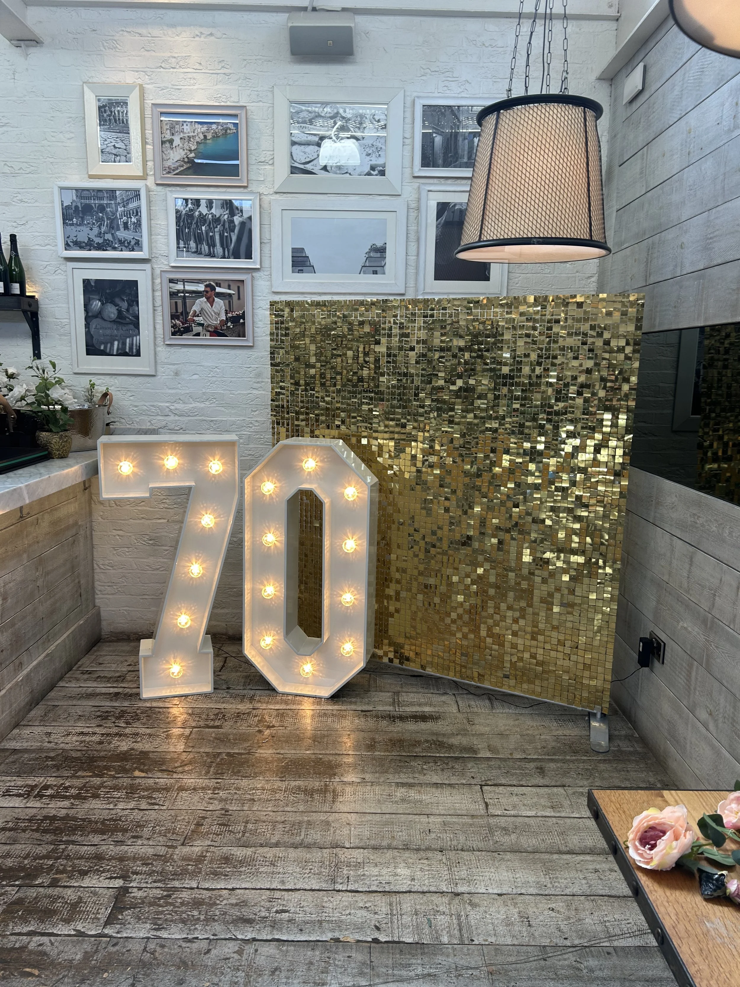 Decorative interior space with a large illuminated '70' sign, a gold mosaic wall, framed photographs on a white brick wall, a hanging lamp, and a table with flowers.