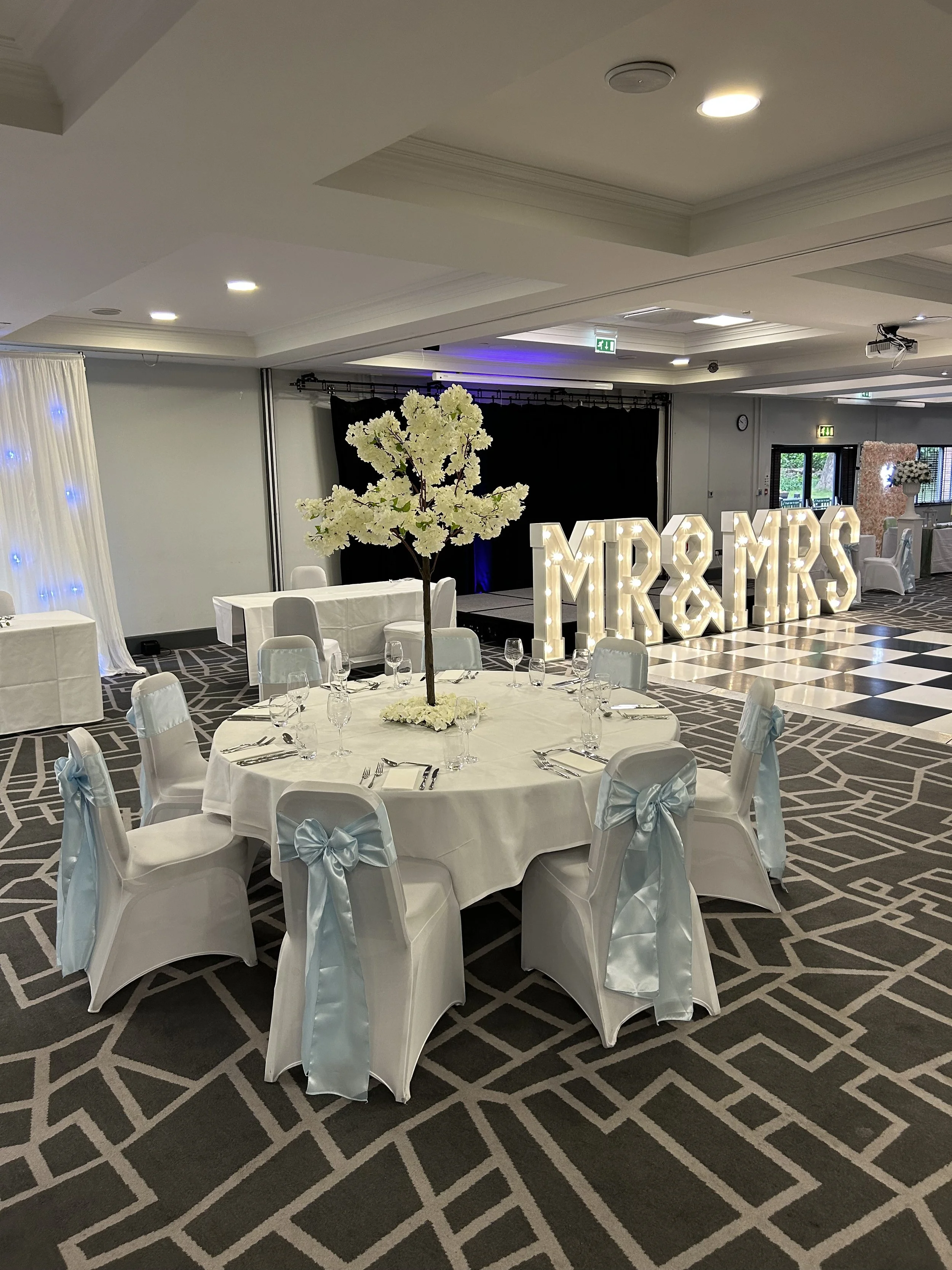 Elegant indoor event venue decorated with white table settings, a centerpiece of white flowers on a round table, and a large illuminated sign with the initials \