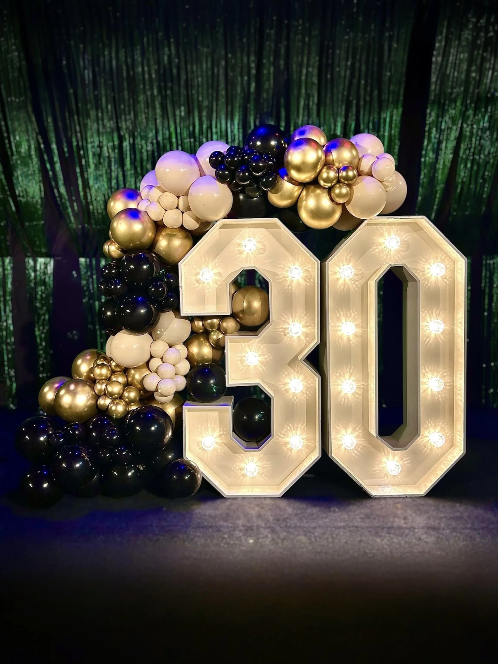 Large illuminated number 30 decorated with black, white, gold, and purple balloons, set against a backdrop of a green forest scene.