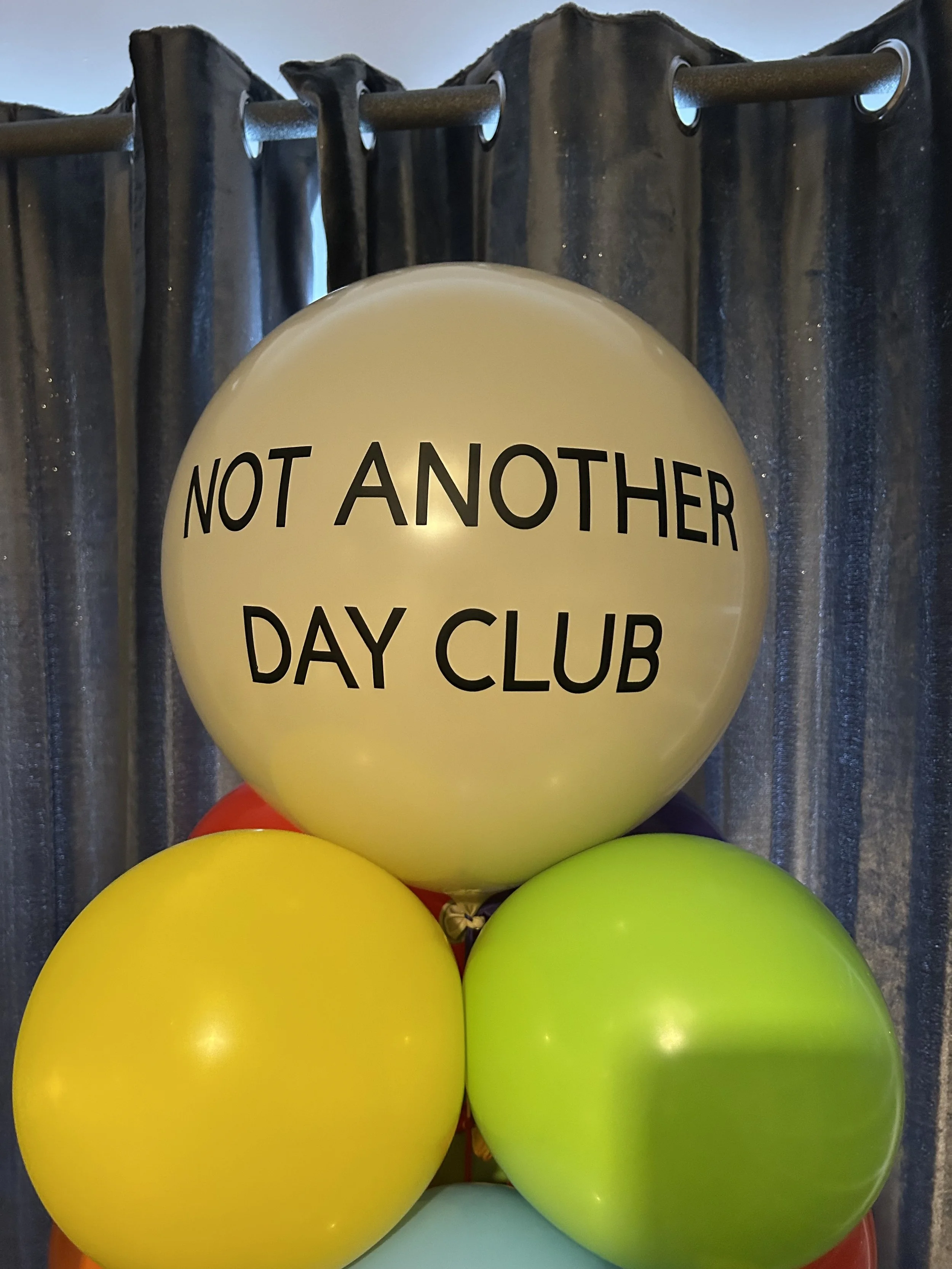 A cluster of colorful balloons with the top balloon displaying the message 'Not Another Day Club' in black text, set against a background of blue and silver curtains.