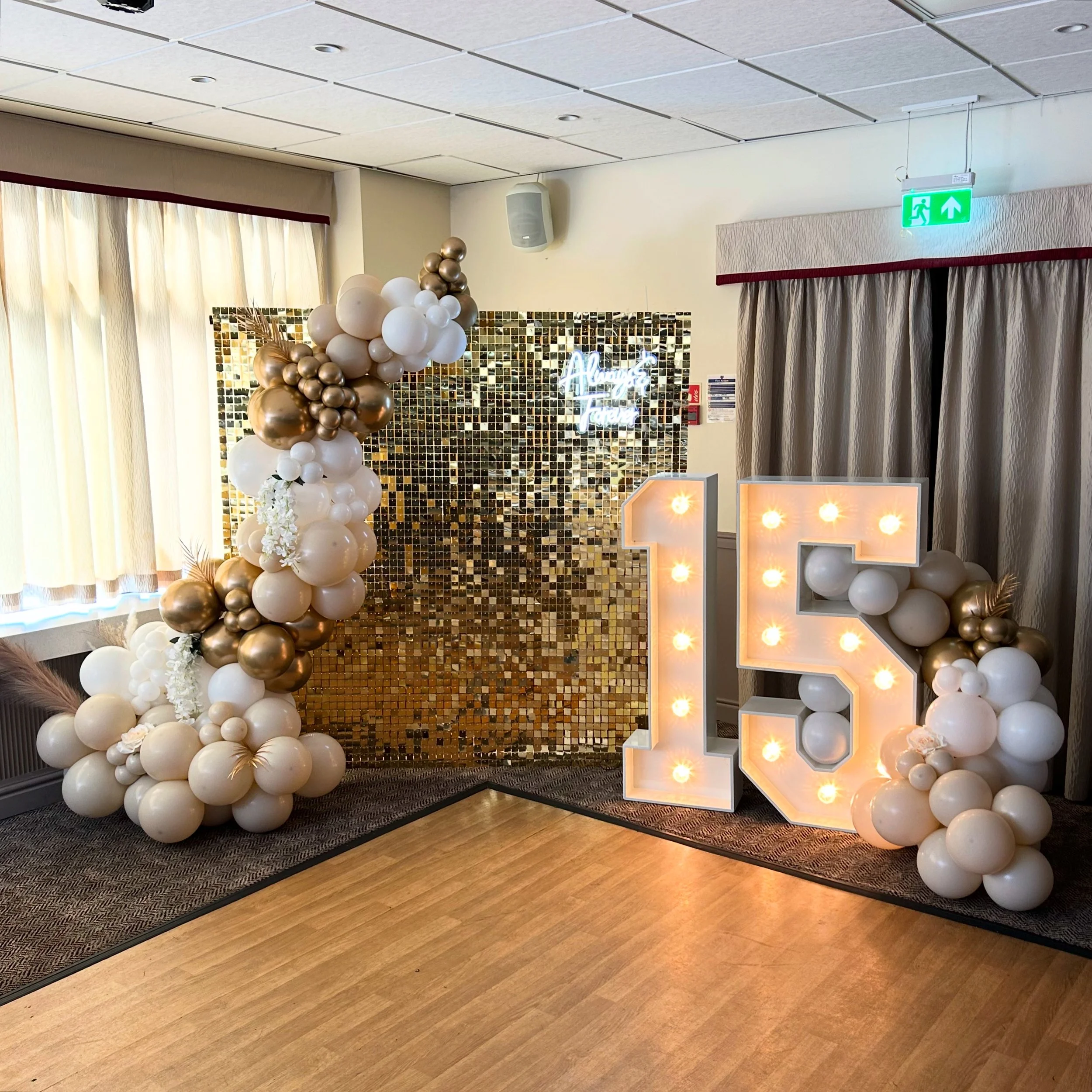 A decorated party backdrop with white, gold, and cream balloons forming an arch and cluster around an illuminated number 15. There is a shiny gold mosaic wall in the background, a neon sign saying 'Always and Forever,' and curtains on one side. The s